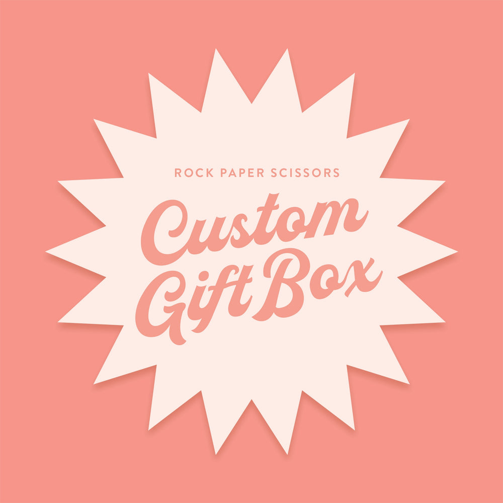On a coral background sits a very light peach starburst. In the starburst it says "ROCK PAPER SCISSORS" in coral, all caps block font. It also says "Custom Gift Box" in coral, thick script font.