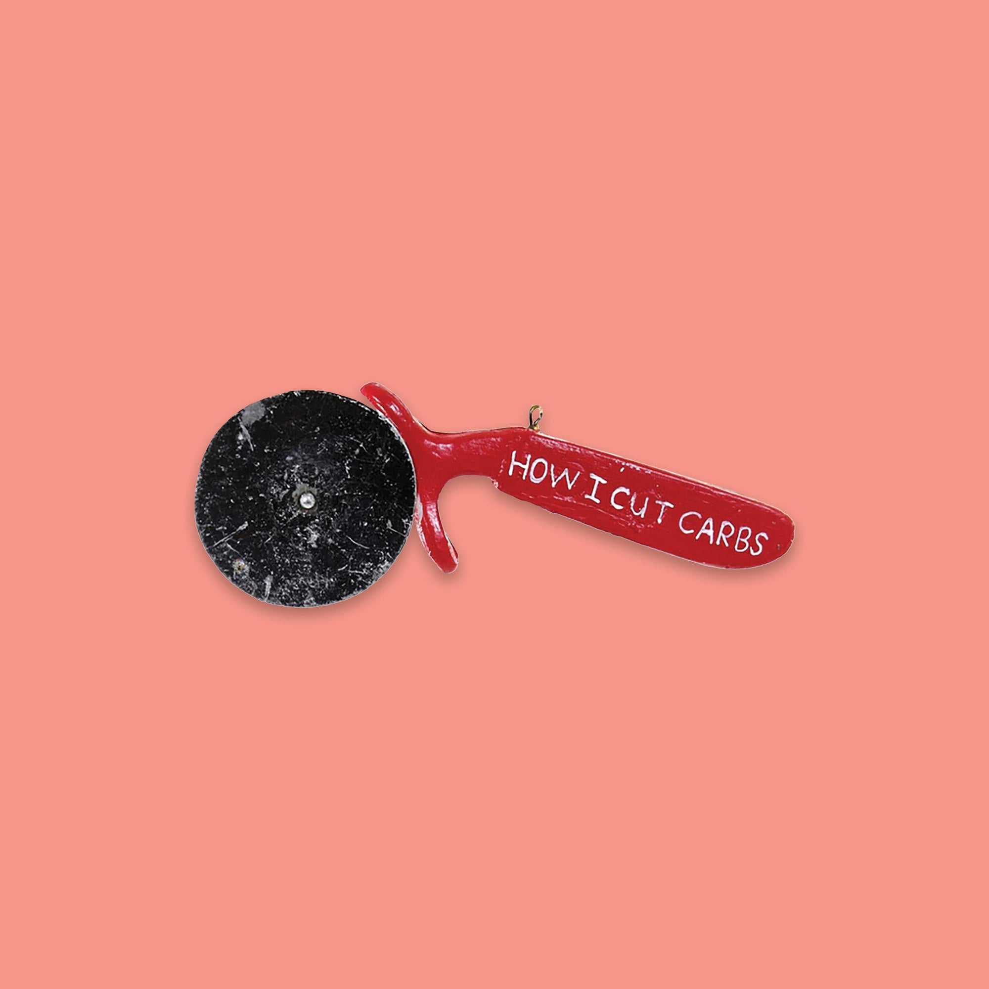 On a coral pink background sits a pizza cutter ornament. This is a glass pizza cutter ornament with a red handle and it says "HOW I CUT CARBS" in white lettering.