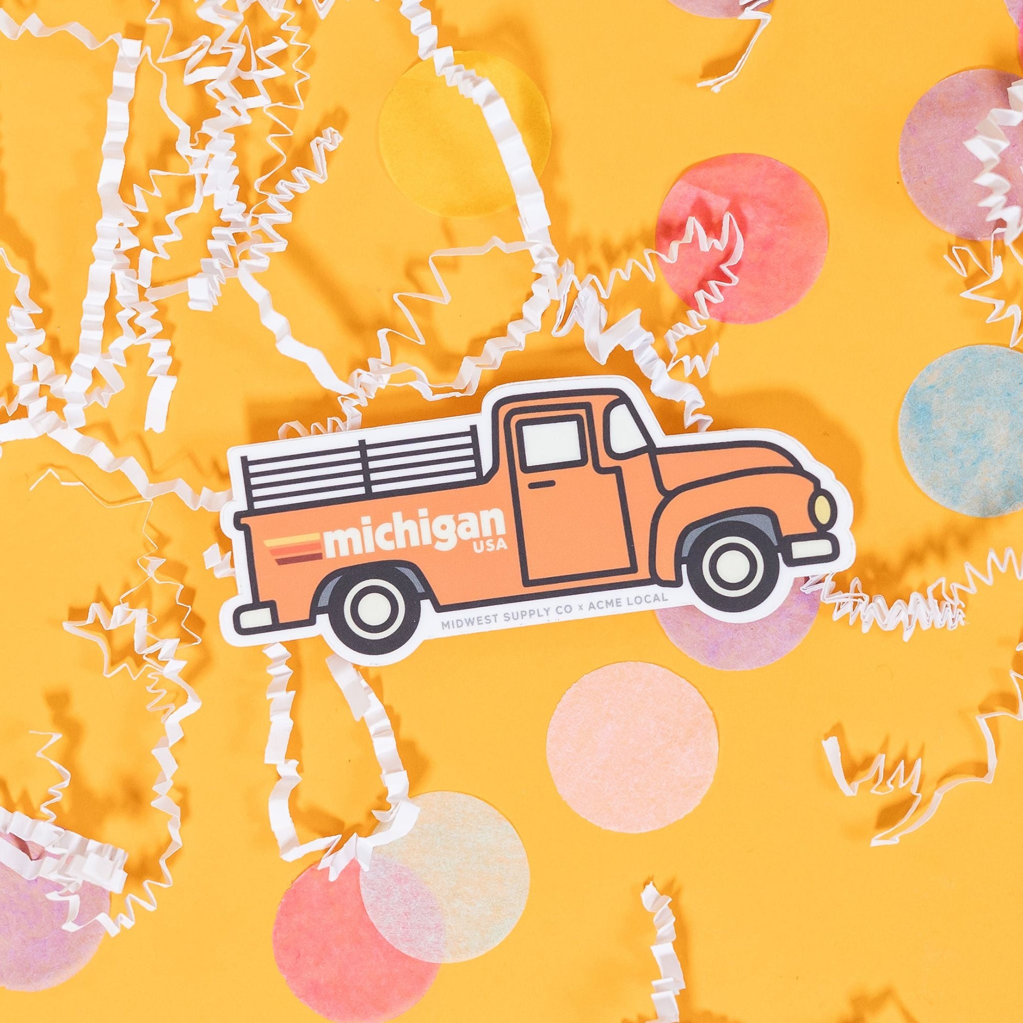 On a sunny mustard background sits a sticker with white crinkle and big, colorful confetti scattered around. This white sticker has an illustration of a vintage red truck with black outlines and it says "michigan USA" in white block lettering with three stripes. The stripes colors are yellow, coral, and maroon. 2"-3"