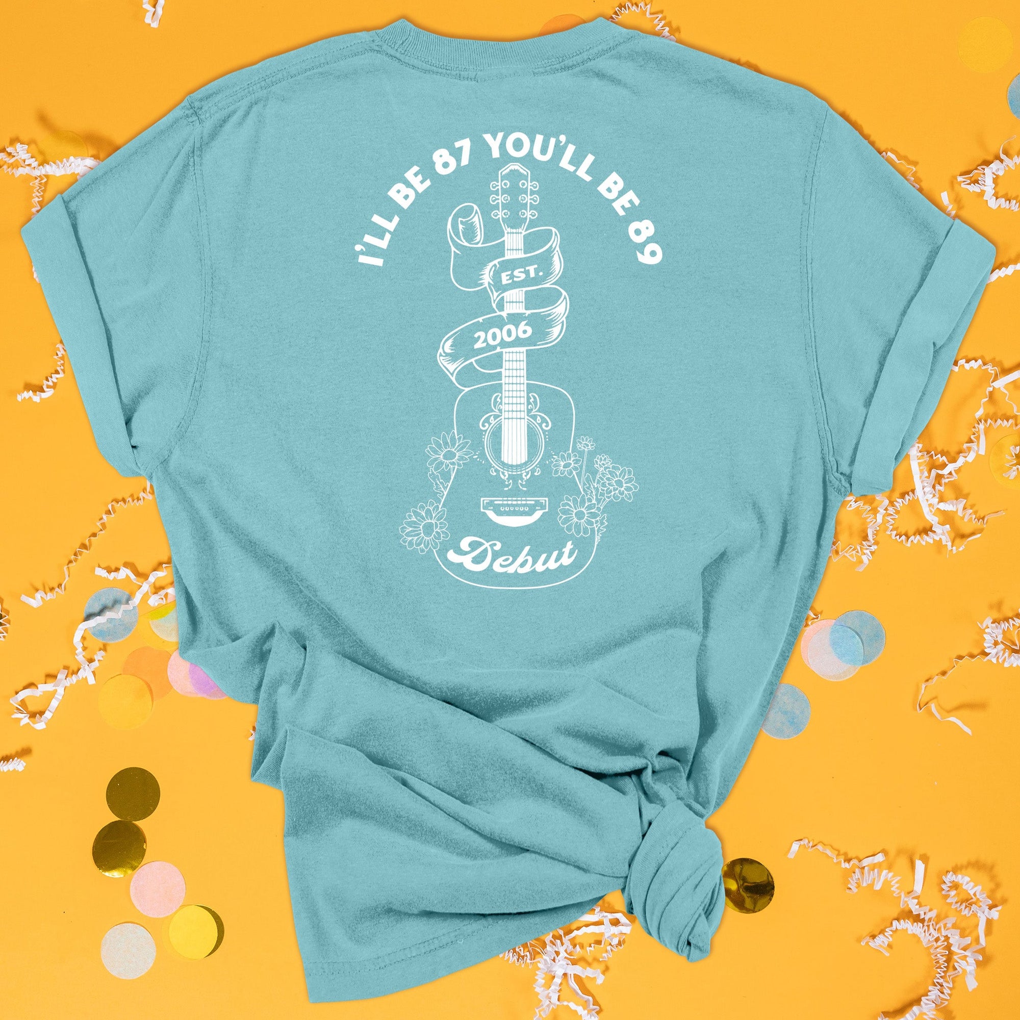 On a sunny mustard background sits the back of a t-shirt with white crinkle and big, colorful confetti scattered around. This Taylor Swift Inspired Reputation tee is aqua with white lettering and illustration. There is a guitar with flowers and a ribbon wrapped around the top of it. It says "I'LL BE 87 YOU'LL BE 89" at the top. In the ribbon it says "EST. 2006" and on the bottom of the guitar it says "Debut."