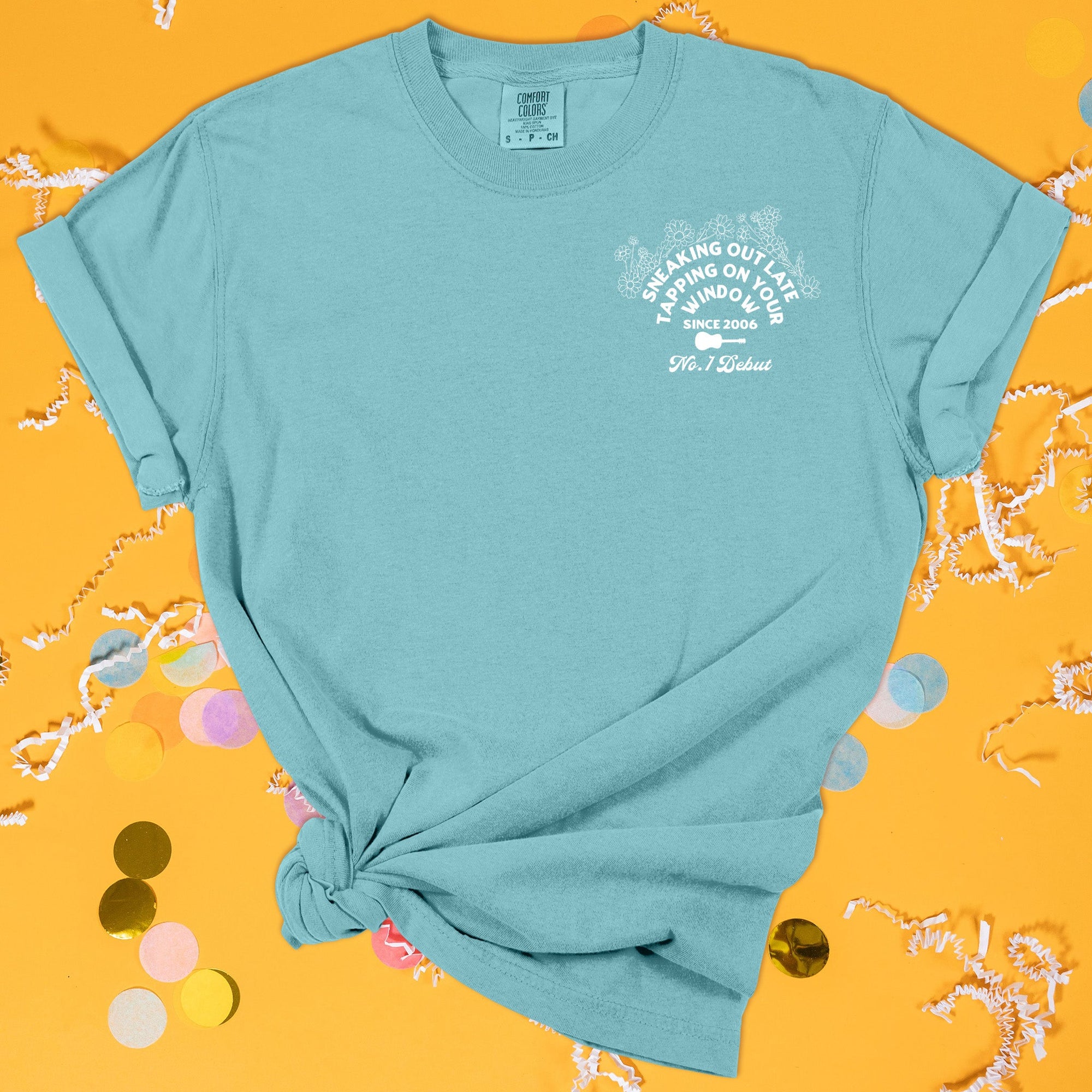 On a sunny mustard background sits the front of a t-shirt with white crinkle and big, colorful confetti scattered around. This Taylor Swift Inspired Reputation tee is aqua with white lettering and illustration. There is a guitar with flowers and it says "SNEAKING OUT LATE TAPPING ON YOUR WINDOW SINCE 2006" and "No. 1 Debut."