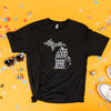 On a sunny mustard background sits a t-shirt with white crinkle and big, colorful confetti scattered around. There are mini disco balls, sunglasses, and a pair of white sneakers. This heathered charcoal grey tee features an illustration of a beer with hops and hand lettered "DRINK GOOD MICHIGAN BEER" in white.