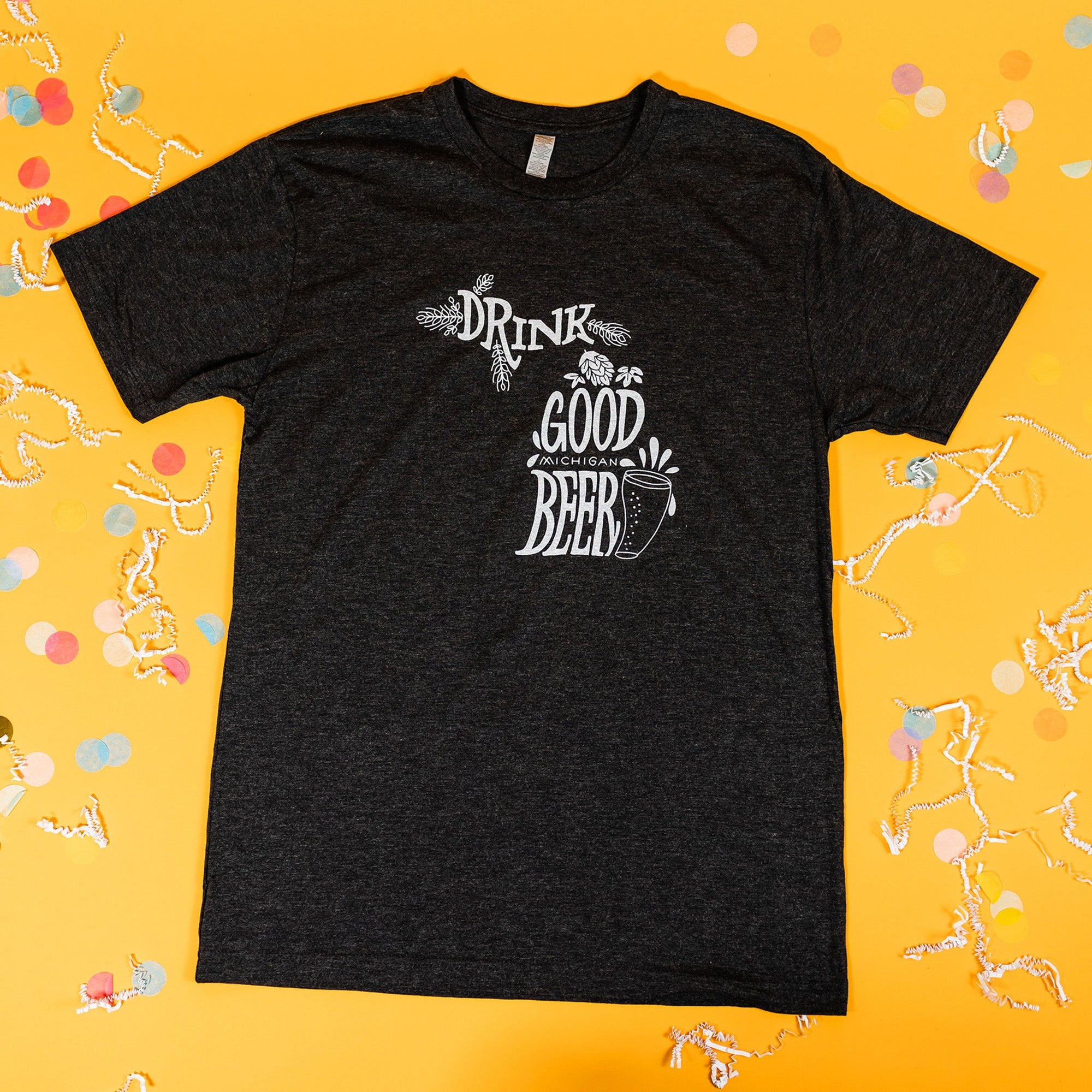 On a sunny mustard background sits a t-shirt with white crinkle and big, colorful confetti scattered around. There are mini disco balls, sunglasses, and a pair of white sneakers. This heathered charcoal grey tee features an illustration of a beer with hops and hand lettered "DRINK GOOD MICHIGAN BEER" in white.