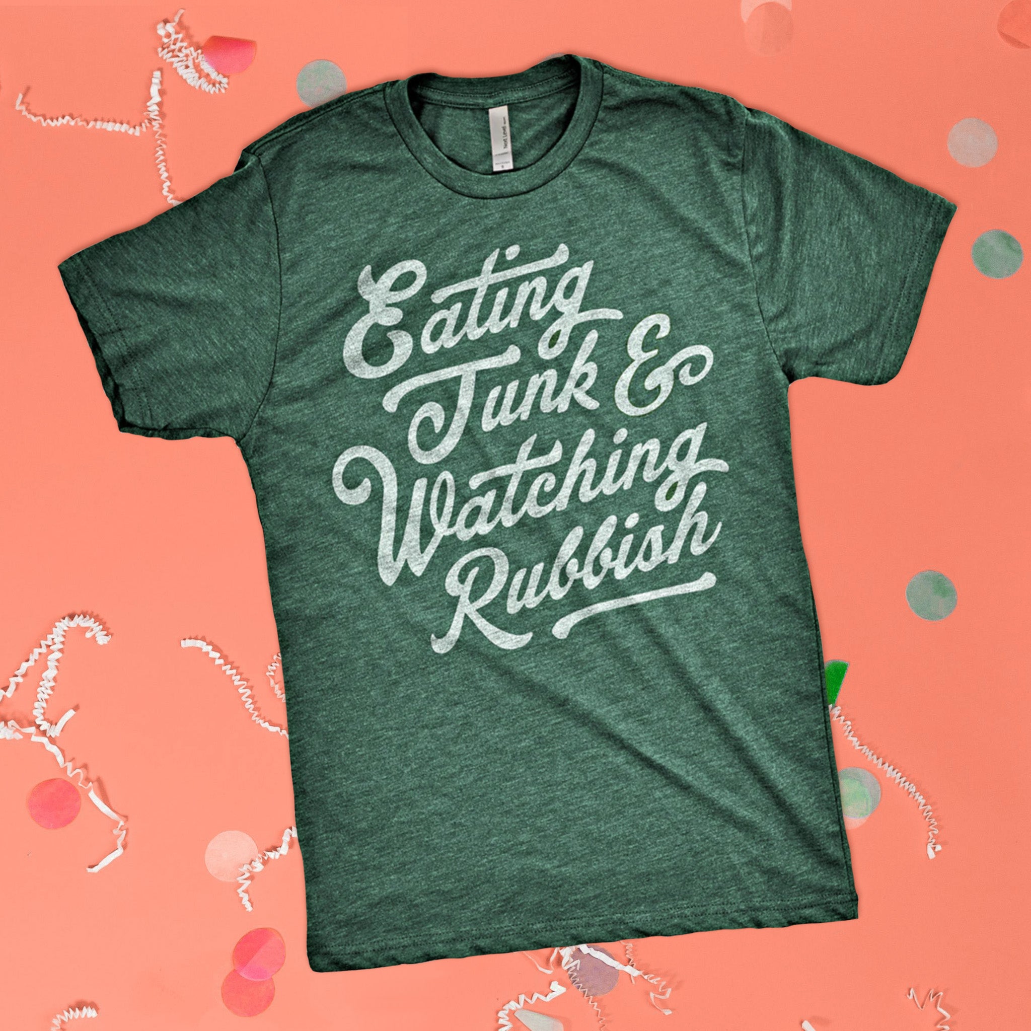 On a pink coral background sits the front of a t-shirt with white crinkle and big, colorful confetti scattered around. This Home Alone Inspired crewneck heathered dark green t-shirt has white script lettering and it says "Eating Junk & Watching Rubbish."