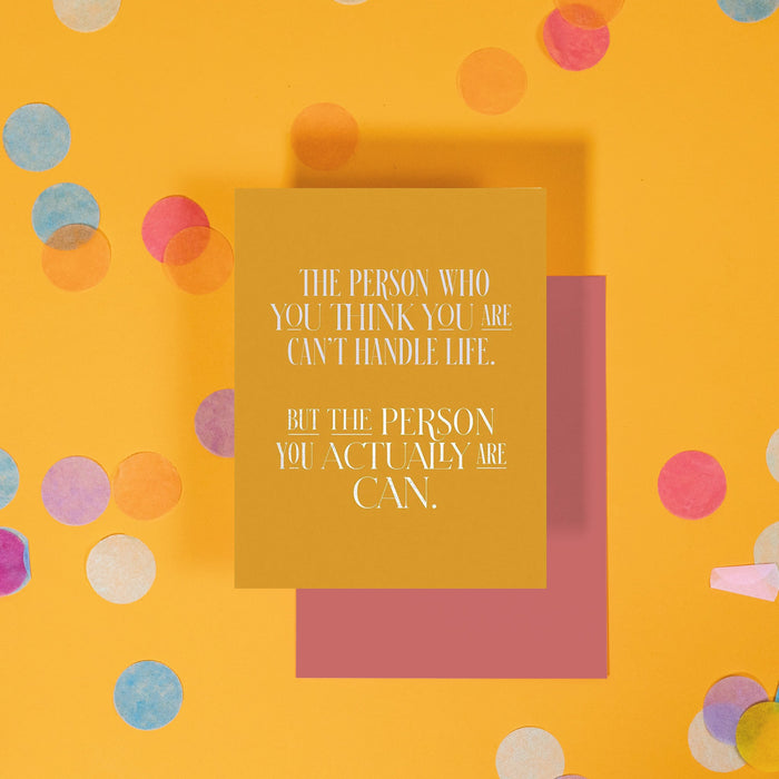 On a sunny mustard background is a greeting card and envelope with big, colorful confetti scattered around. The golden mustard greeting card says "The person who you think you are can't handle life. But the person you actually are can." in a white, all caps, serif font. The marsala colored envelope sits under the card.