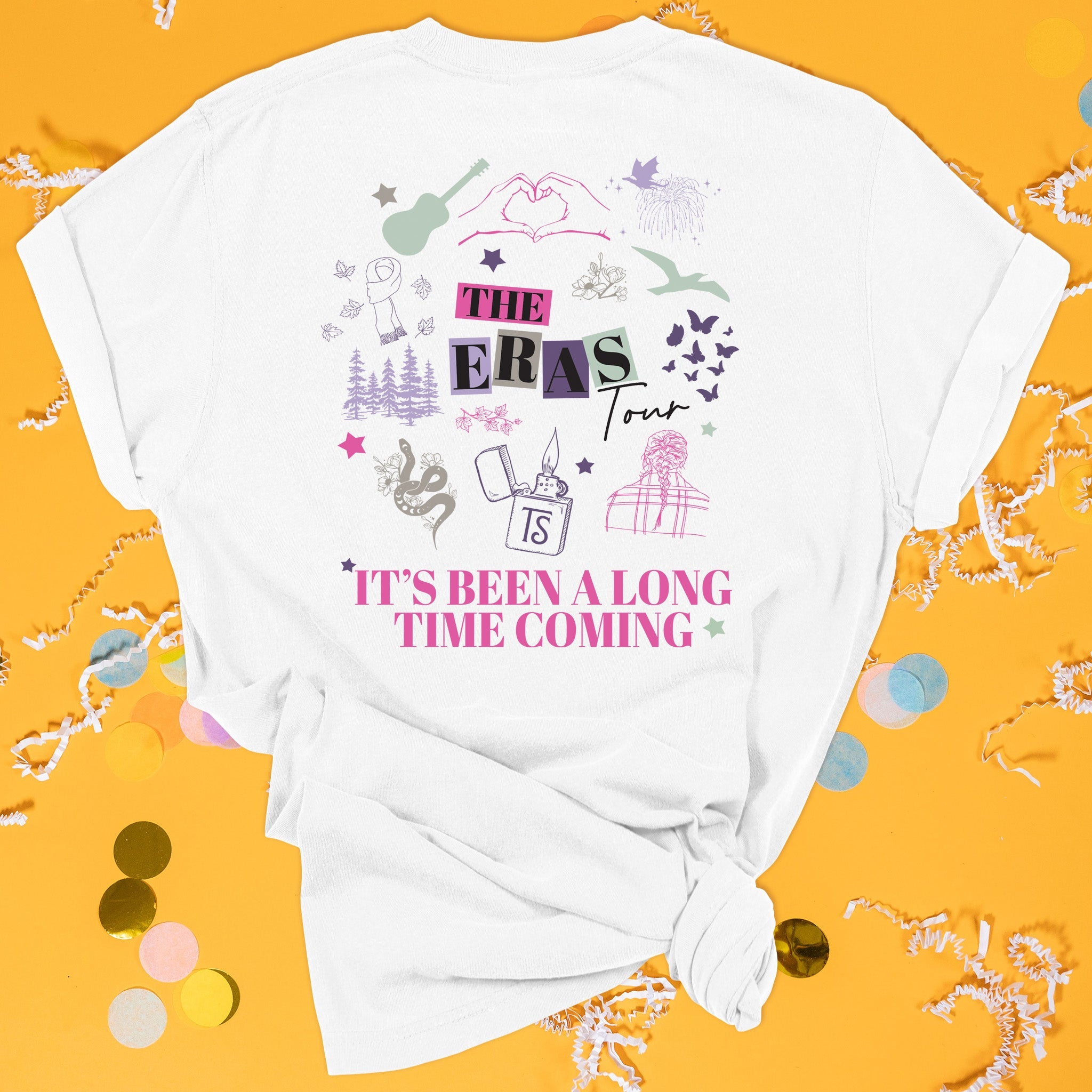 On a sunny mustard background sits the back of a t-shirt with white crinkle and big, colorful confetti scattered around. This Taylor Swift Inspired Eras Tour tee is white with hot pink lettering and an illustration of all the eras icons in colors of hot pink, dark purple, lavender, sage, and khaki. It says "IT'S BEEN A LONG TIME COMING" in all caps and "THE EARS Tour" black lettering with colorful blocks. 