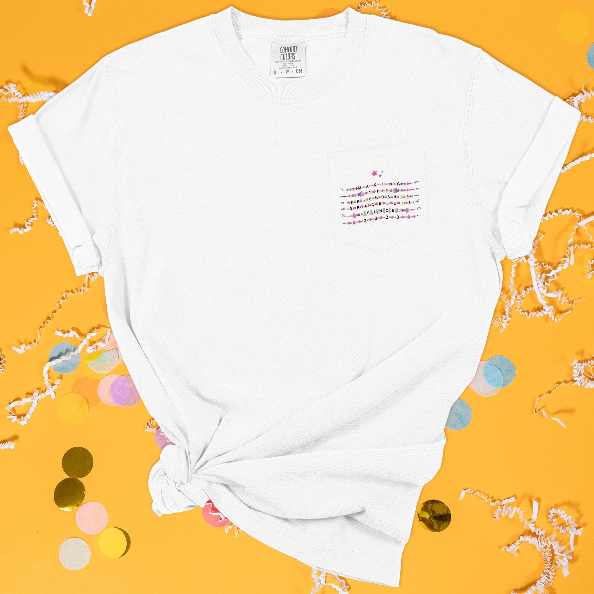 On a sunny mustard background sits the front of a t-shirt with white crinkle and big, colorful confetti scattered around. This Taylor Swift Inspired Eras Tour tee is white with a pocket over the left chest area and it has an illustration of six friendship bracelets in colors of hot pink, dark purple, lavender, sage, and khaki. Each bracelet has a word and it says "MAKING THE FRIENDSHIP BRACELETS SINCE 2023" in all caps black lettering. There are three stars above the bracelets in hot pink, purple, and sage.