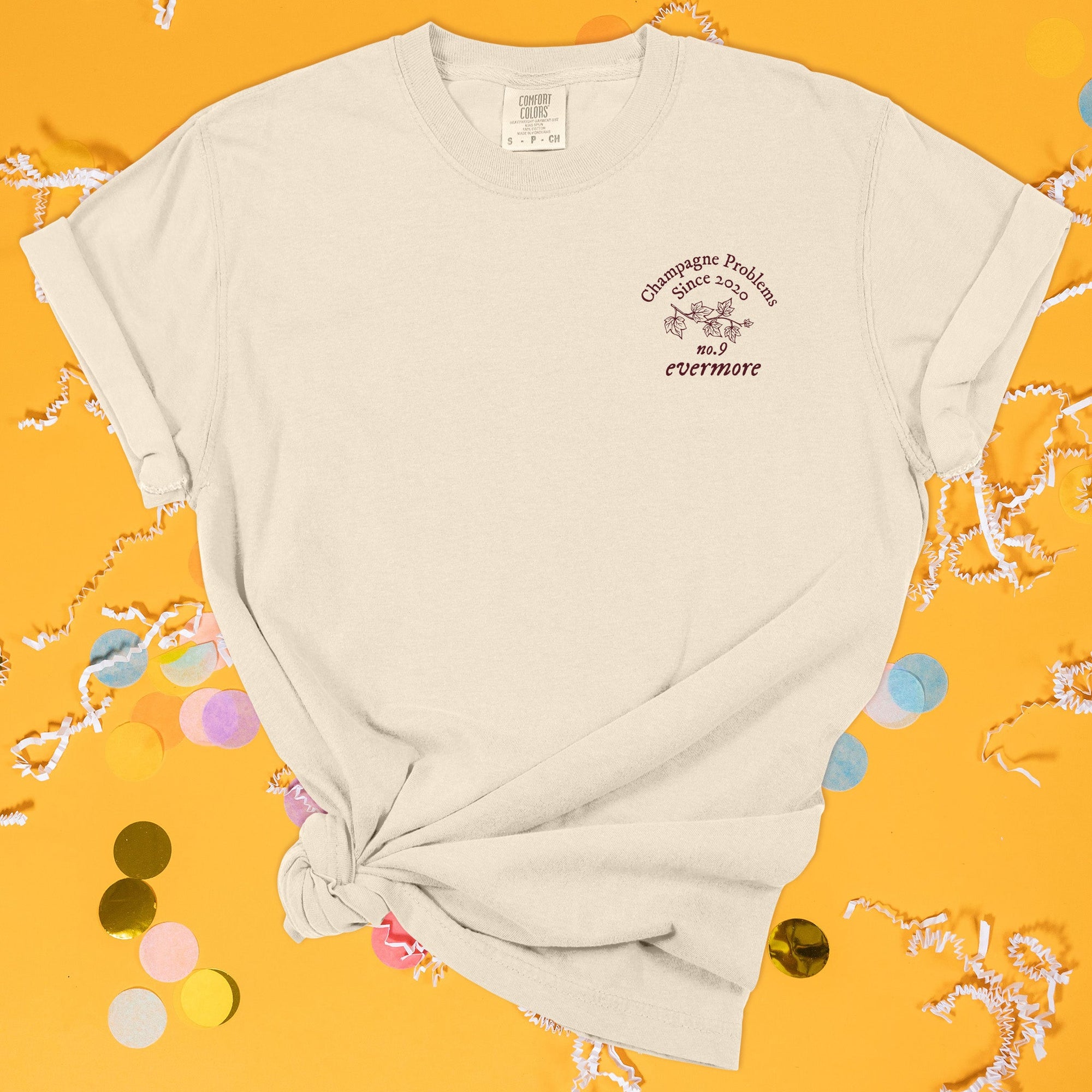 On a sunny mustard background sits the front of a t-shirt with white crinkle and big, colorful confetti scattered around. This Taylor Swift Inspired Reputation tee is ivory with maroon lettering and illustration over the left chest area. It has an illustration of fall leaves on a branch and it says "Champagne Problems Since 2020, No.9 evermore."