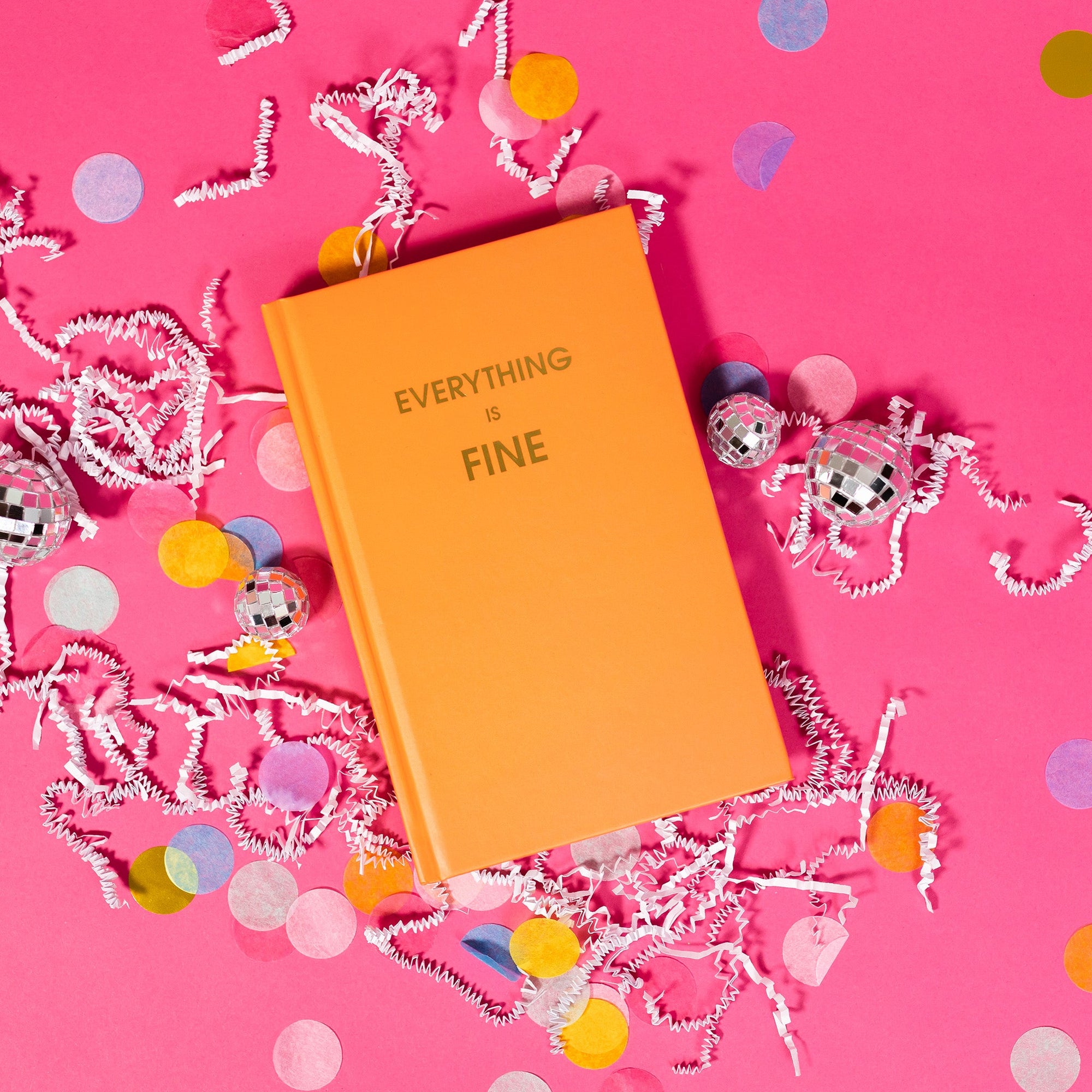 On a hot pink background sits a journal with white crinkle and big, colorful confetti scattered around. There are mini disco balls. This orange journal says "EVERYTHING IS FINE" in gold, all caps block font.