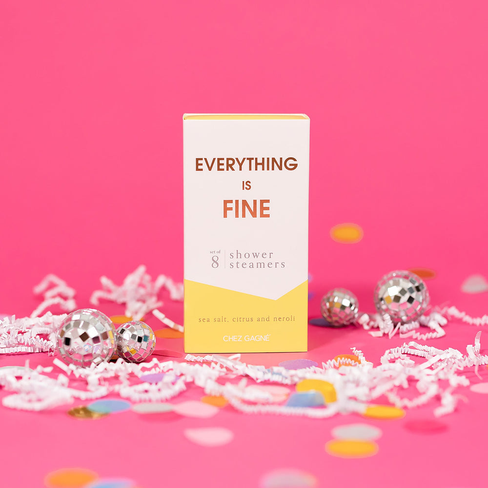 On a hot pink background sits a box with white crinkle and big, colorful confetti scattered around. There are mini disco balls. This picture is a close-up of a white and yellow package that says "EVERYTHING IS FINE" in gold foil, all caps block lettering. Under it says "set of 8" and " shower steamers" in grey, lowercase serif font. At the bottom it says "sea salt citrus and neroli" in gold foil, lower case serif font.