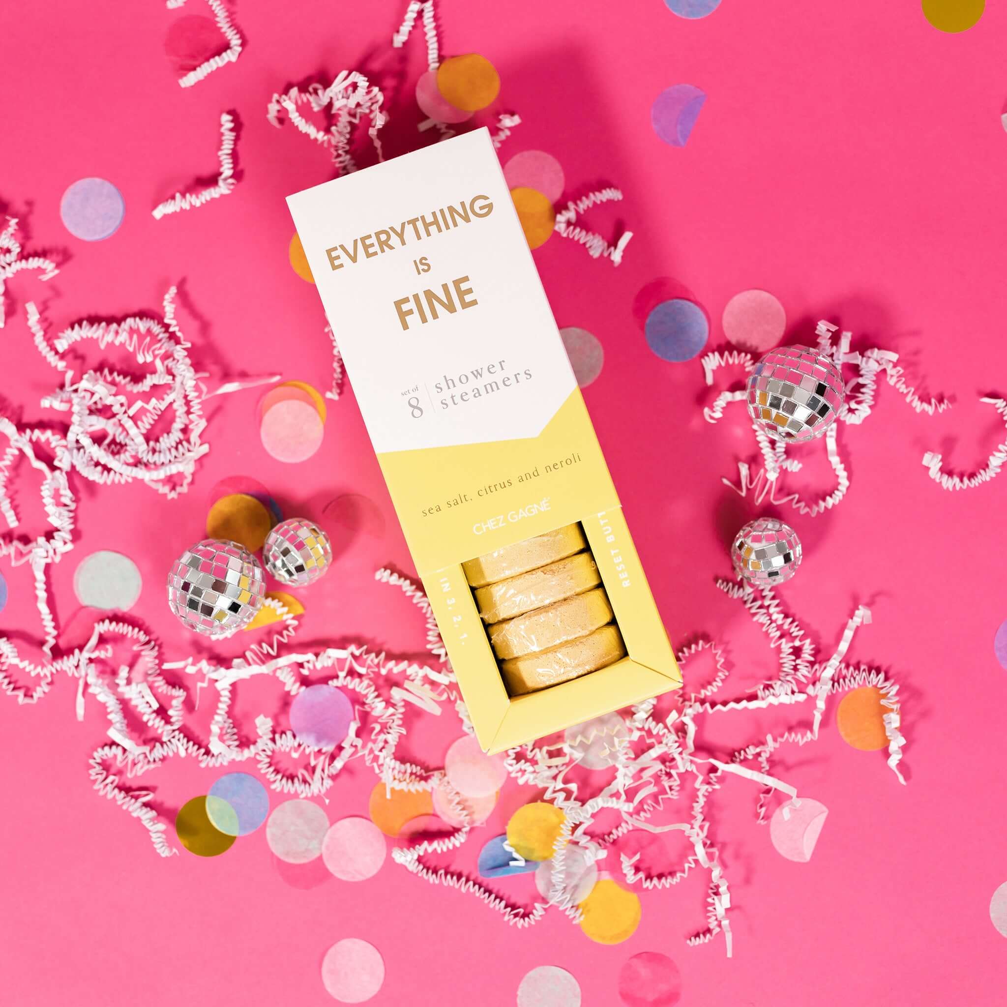 On a hot pink background sits an opened box with white crinkle and big, colorful confetti scattered around. There are mini disco balls. This picture is a close-up of a white and yellow package that says "EVERYTHING IS FINE" in gold foil, all caps block lettering. Under it says "set of 8" and " shower steamers" in grey, lowercase serif font. At the bottom it says "sea salt citrus and neroli" in gold foil, lower case serif font. The box is opened to reveal the yellow shower steamers in it.