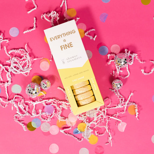On a hot pink background sits an opened box with white crinkle and big, colorful confetti scattered around. There are mini disco balls. This picture is a close-up of a white and yellow package that says "EVERYTHING IS FINE" in gold foil, all caps block lettering. Under it says "set of 8" and " shower steamers" in grey, lowercase serif font. At the bottom it says "sea salt citrus and neroli" in gold foil, lower case serif font. The box is opened to reveal the yellow shower steamers in it.