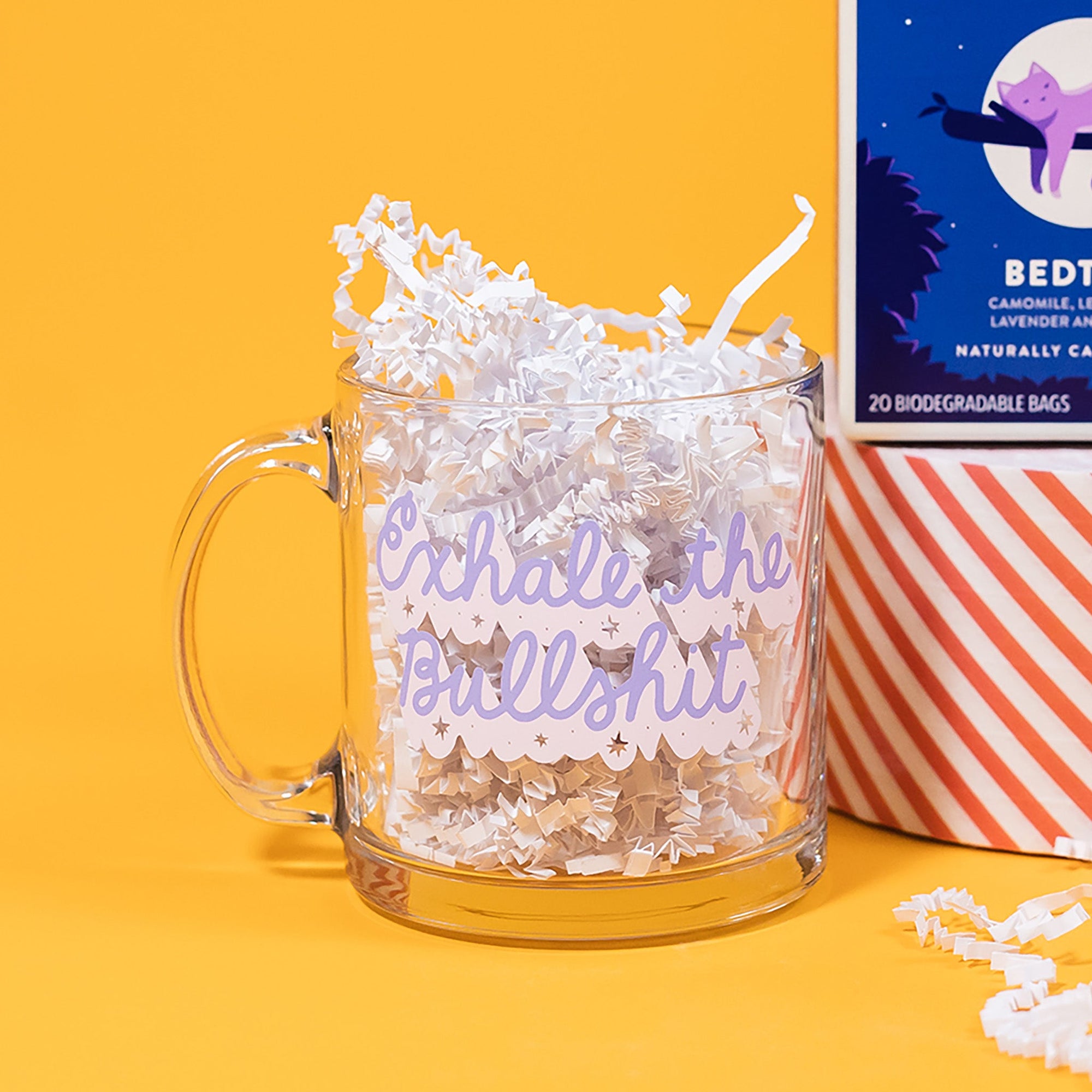 On a sunny mustard background sits a mug with white crinkle in it. This clear mug says "Exhale the Bullshit" in a dark lavender, handwritten script lettering. It has a light lavender dropshadow and stars and dots around the wording.