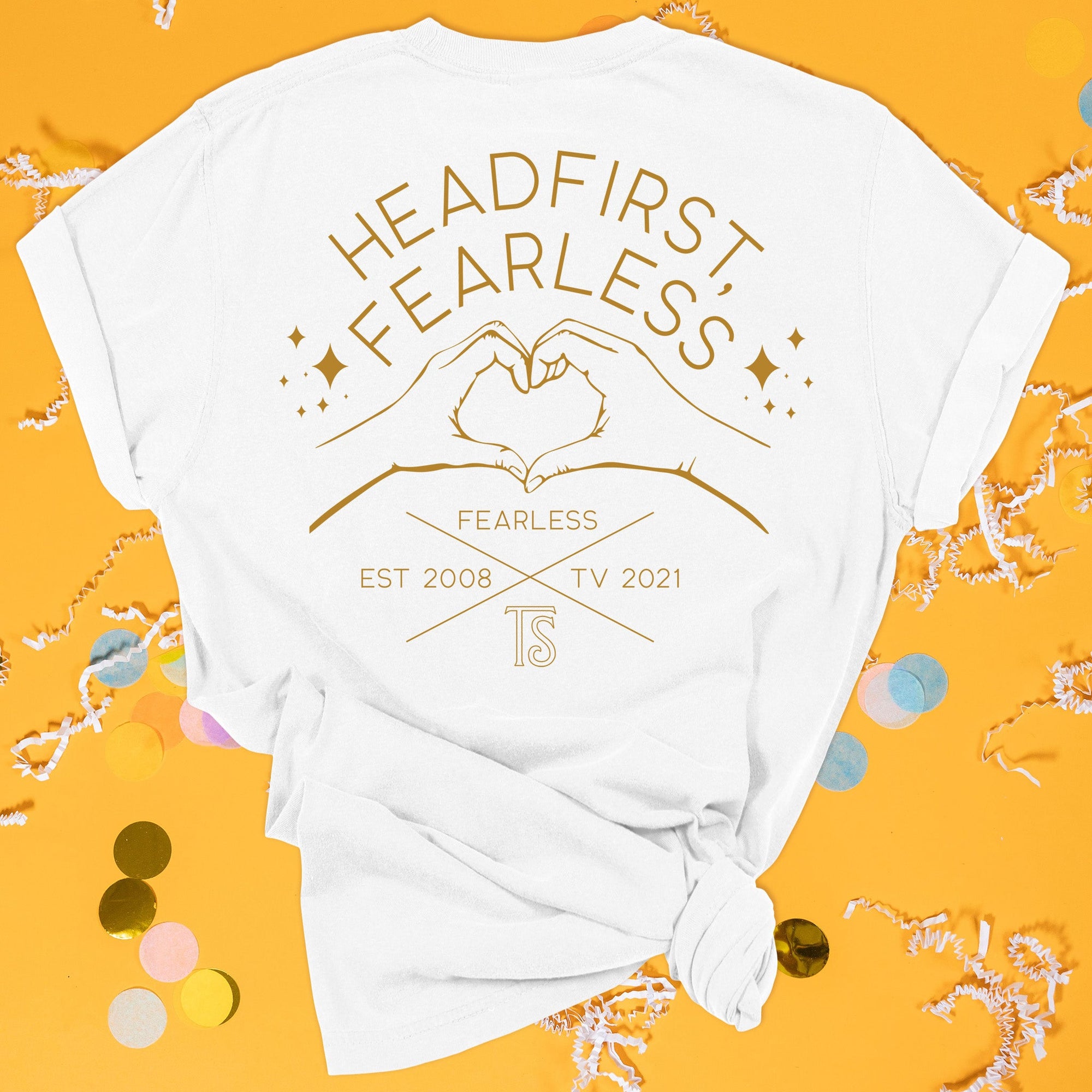 On a sunny mustard background sits the back of a t-shirt with white crinkle and big, colorful confetti scattered around. This Taylor Swift Inspired Reputation tee is white with gold lettering and illustration. There is a hands heart with sparkle stars and it says "HEADFIRST, FEARLESS" in all caps. It also says "FEARLESS, EST 2008, TV 2021, TS" in all caps. 