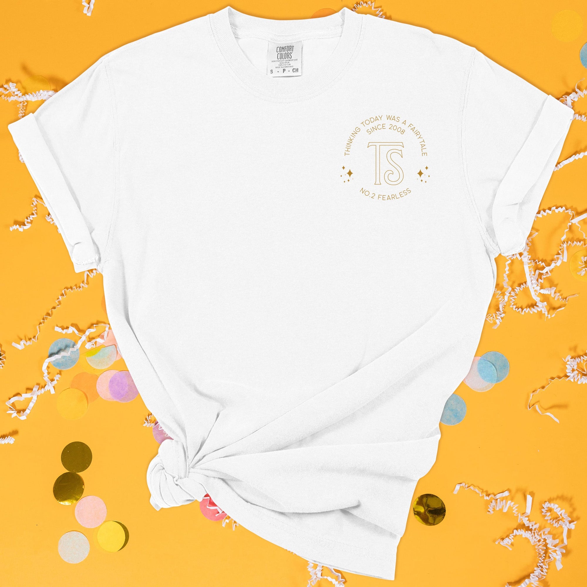 On a sunny mustard background sits the front of a t-shirt with white crinkle and big, colorful confetti scattered around. This Taylor Swift Inspired Reputation tee is white with gold lettering. There is a "TS" with shimmer sparkles over the left chest area and around it says "THINKING TODAY WAS A FAIRYTALE SINCE 2008, NO. 2 FEARLESS" in all caps.