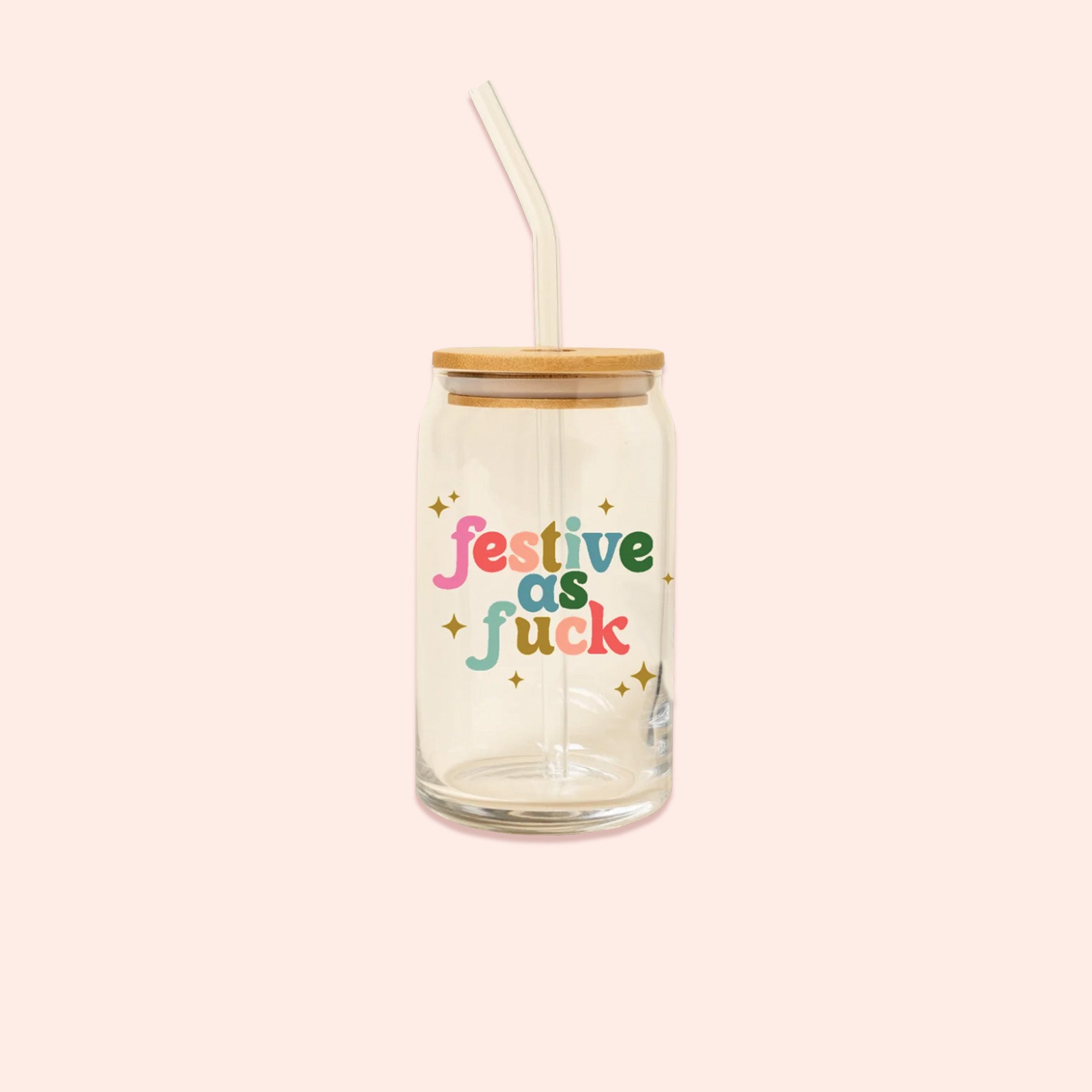 On a light pink background sits a can glass . This can glass with a lid and straw has colorful lettering and gold stars. It says "festive as fuck."