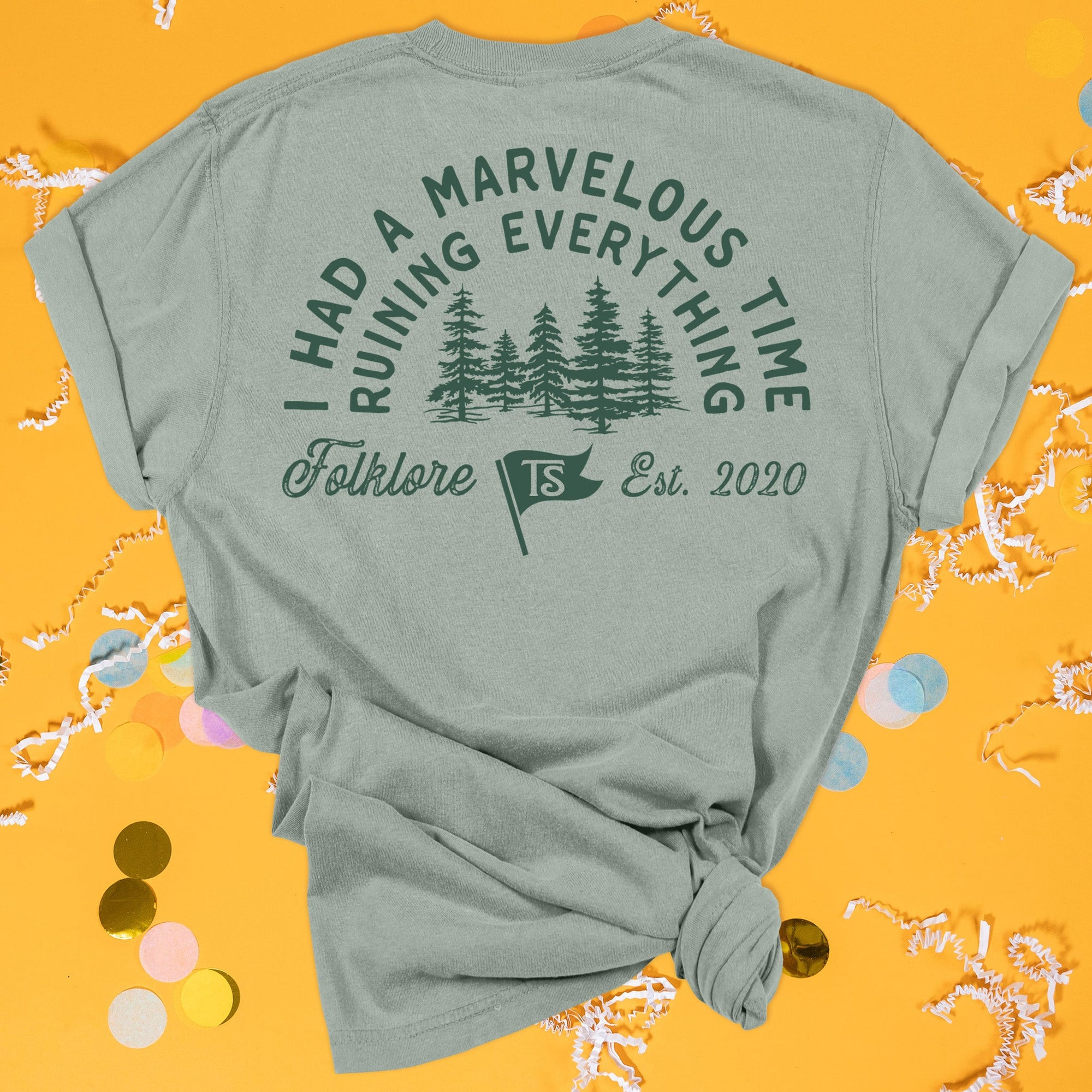 On a sunny mustard background sits the back of a t-shirt with white crinkle and big, colorful confetti scattered around. This Taylor Swift Inspired Reputation tee is dark sage with dark green lettering and an illustration of forest trees. It says "I HAD A MARVELOUS TIME RUINING EVERYTHING" in all caps and "Folklore Est. 2020" in script lettering. There is a flag with "TS" in it.