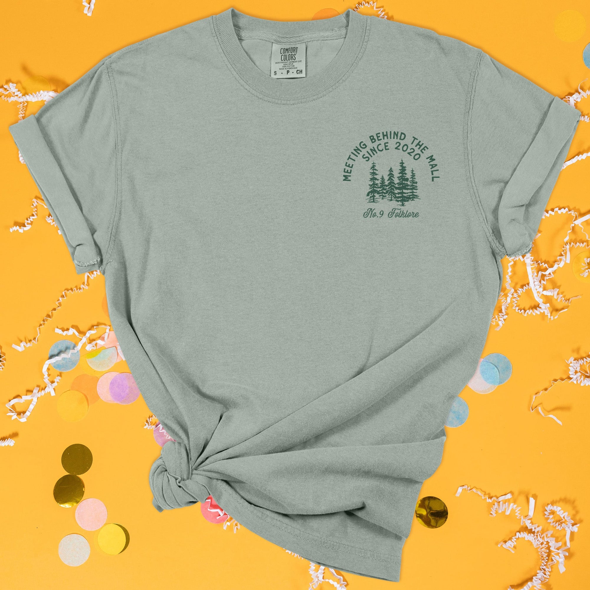 On a sunny mustard background sits the front of a t-shirt with white crinkle and big, colorful confetti scattered around. This Taylor Swift Inspired Reputation tee is dark sage with dark green lettering and an illustration of forest trees over the left chest area. It says "MEETING BEHIND THE MALL SINCE 2020" in all caps and "No.9 Folklore" in script lettering.