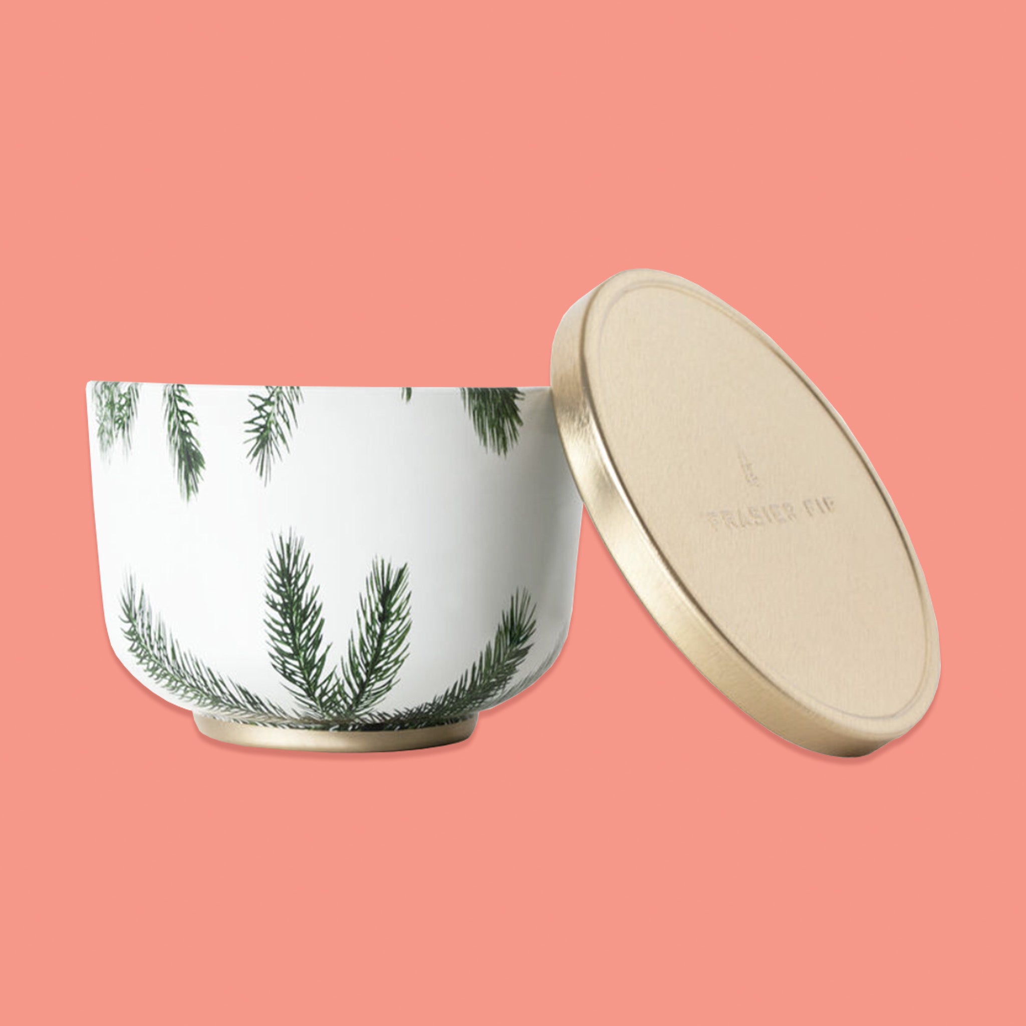 On a pink coral background sits a tin bowl candle that is white with illsutrations of pine branches. Leaning next to it is a gold lid. 