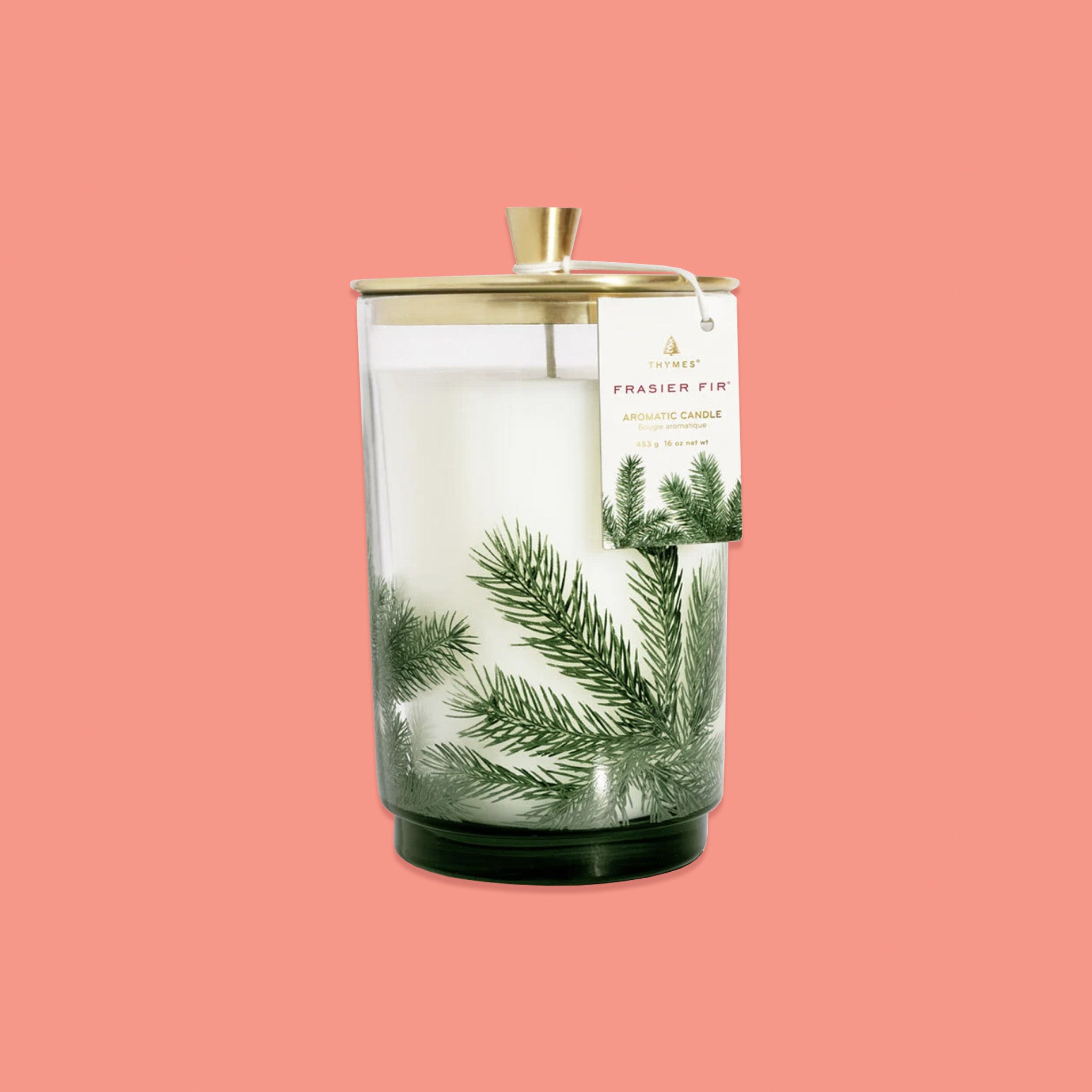 On a pink coral background sits a glass candle with a gold lid. There is an illustration of pine leaves on the glass and a tag hanging off the side. On the tag it says "THYMES FRASIER FIR AROMATIC CANDLE" in gold and red lettering.