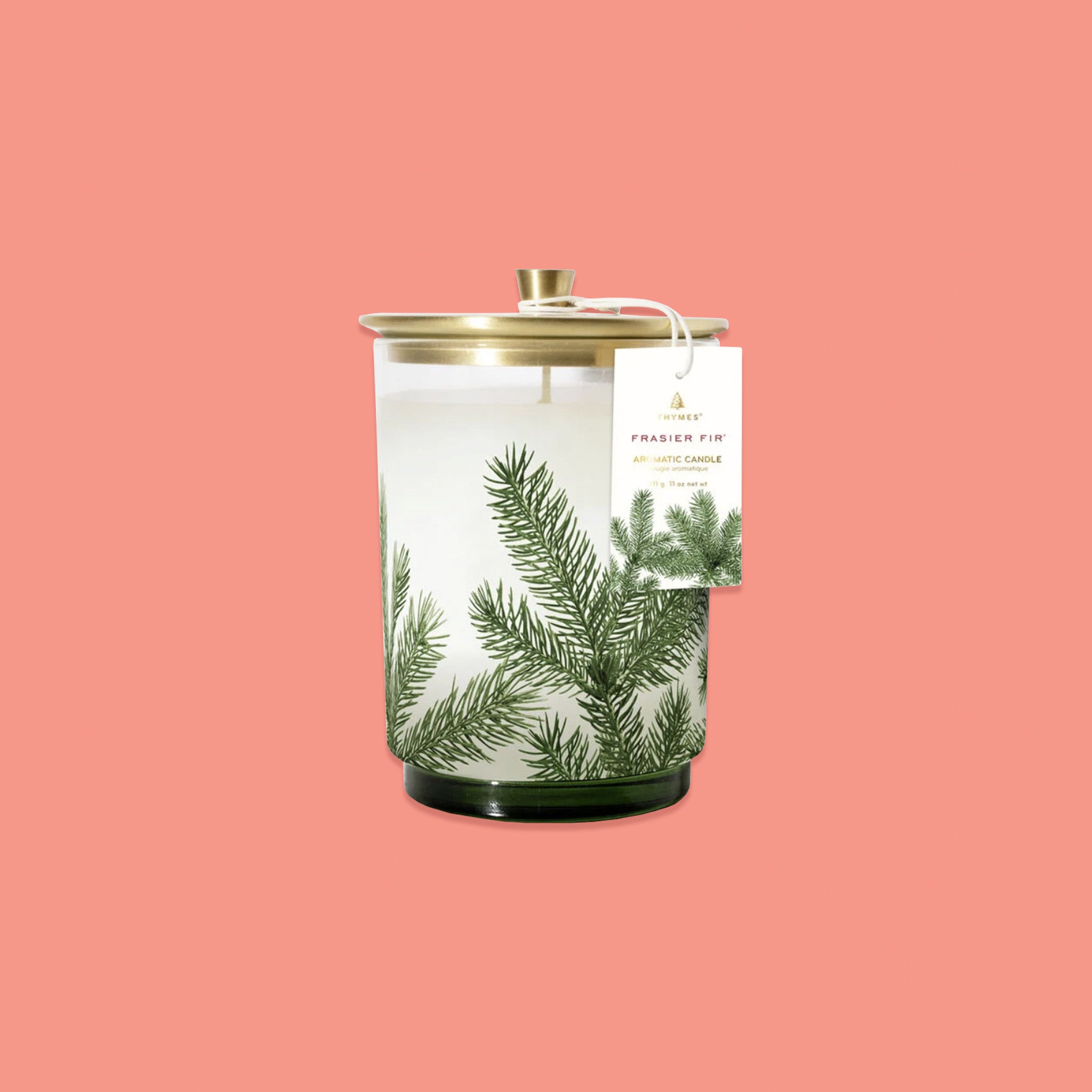 On a pink coral background sits a glass candle with a gold lid. There is an illustration of pine leaves on the glass and a tag hanging off the side. On the tag it says "THYMES FRASIER FIR AROMATIC CANDLE" in gold and red lettering.