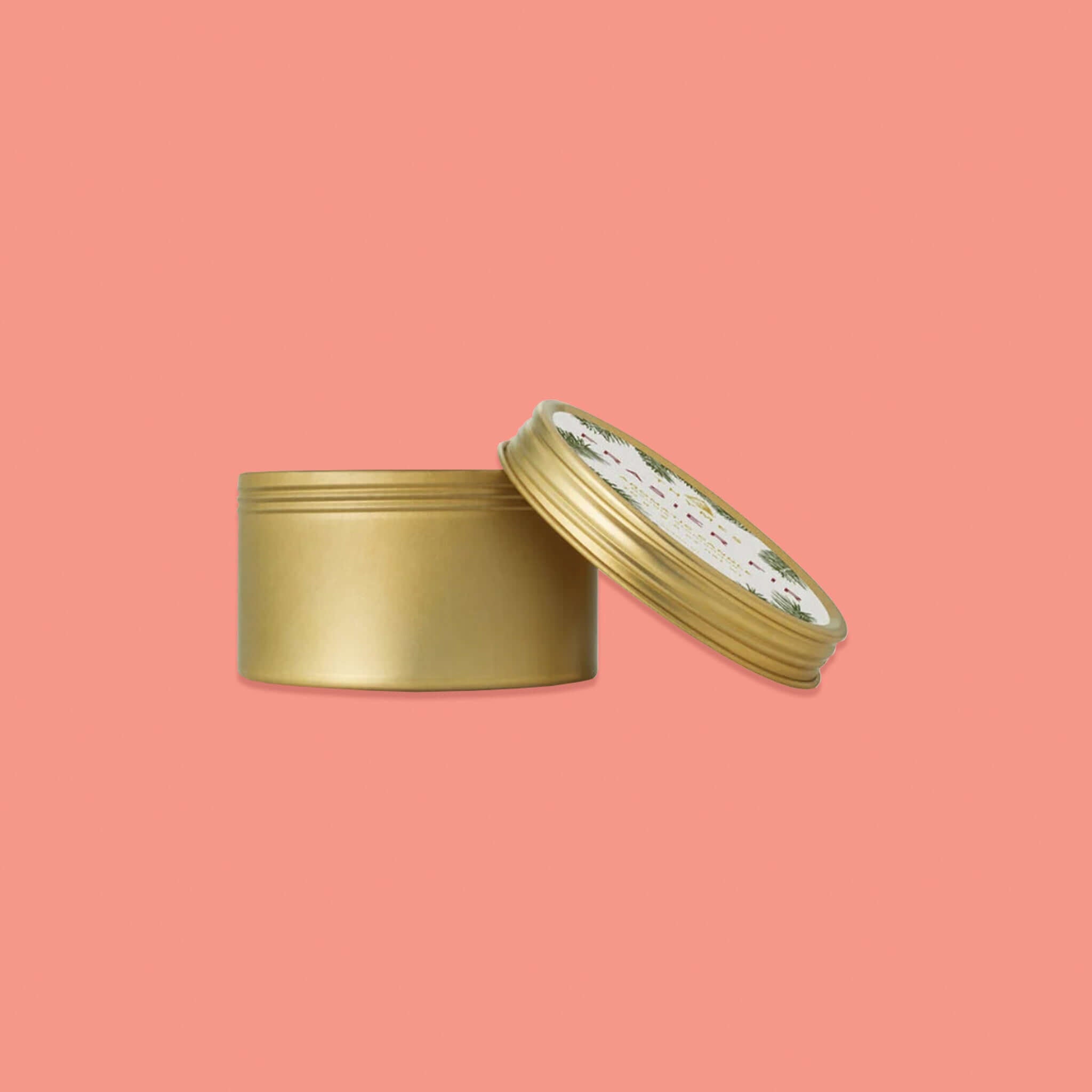 On a pink coral background sits a gold tin travel candle with a gold lid off of the side. There is a label on the lid with illustrations of pine branches. It's says "THYMES FRASIER FIR AROMATIC CANDLE." 70 g  2.5 oz net wt