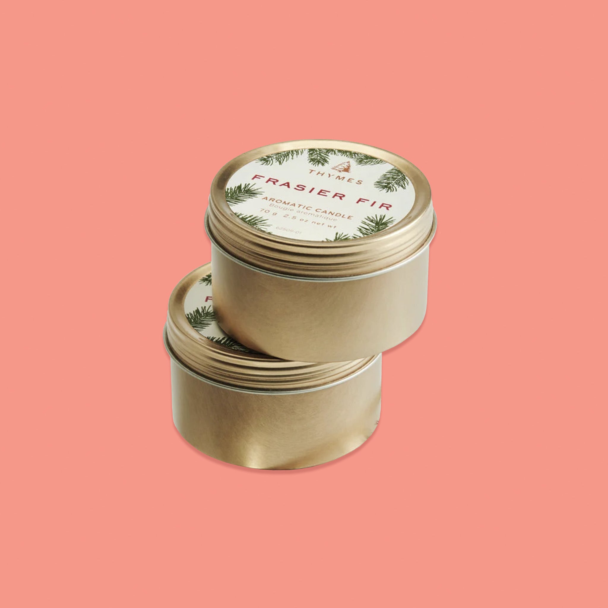 On a pink coral background sits two gold tin travel candles with gold lids stacked on each other. There are labels on the lids with illustrations of pine branches. It's says "THYMES FRASIER FIR AROMATIC CANDLE." 70 g  2.5 oz net wt