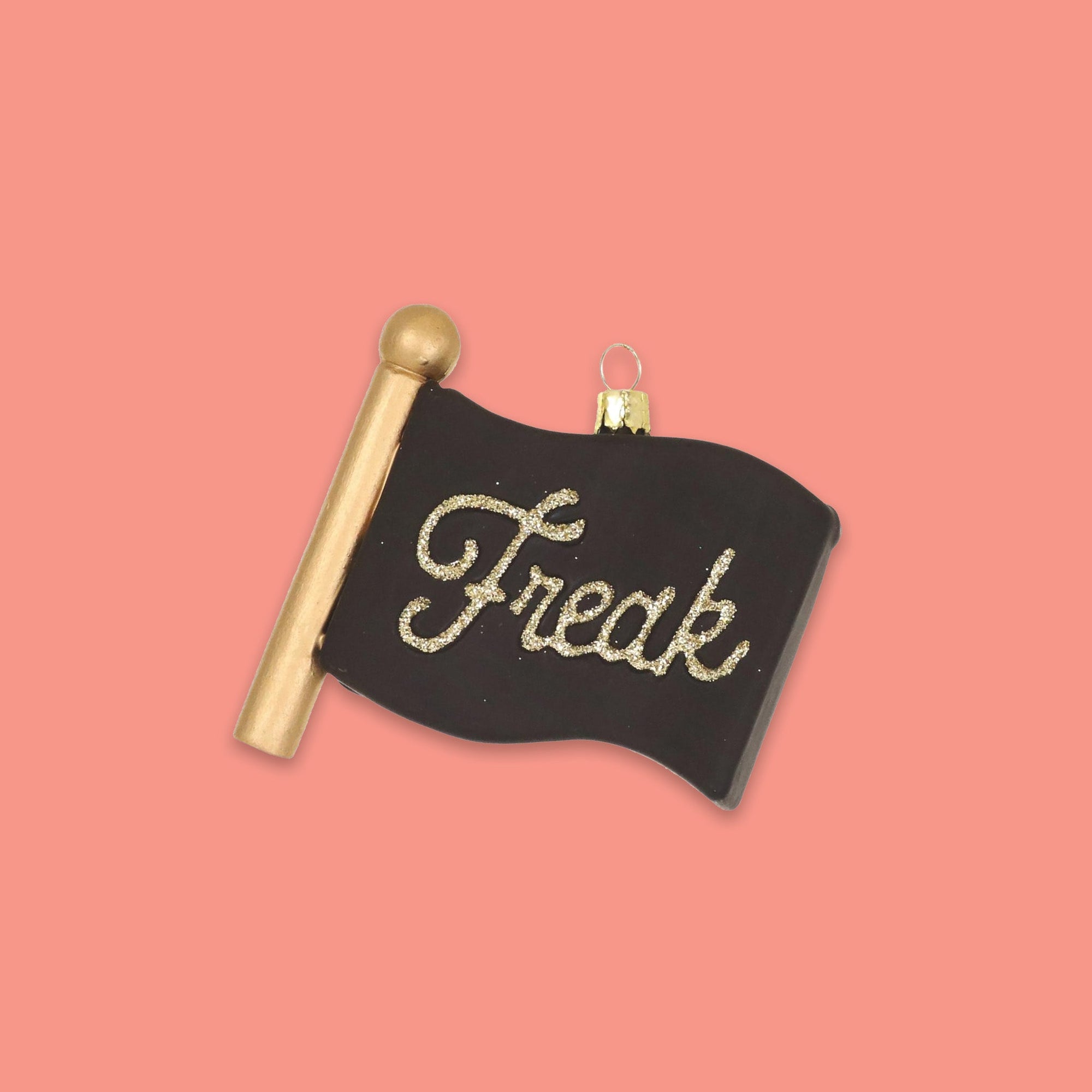 On a coral pink background sits a flag ornament. This black flag and gold pole glass ornament says "Freak" in glitter gold script lettering.