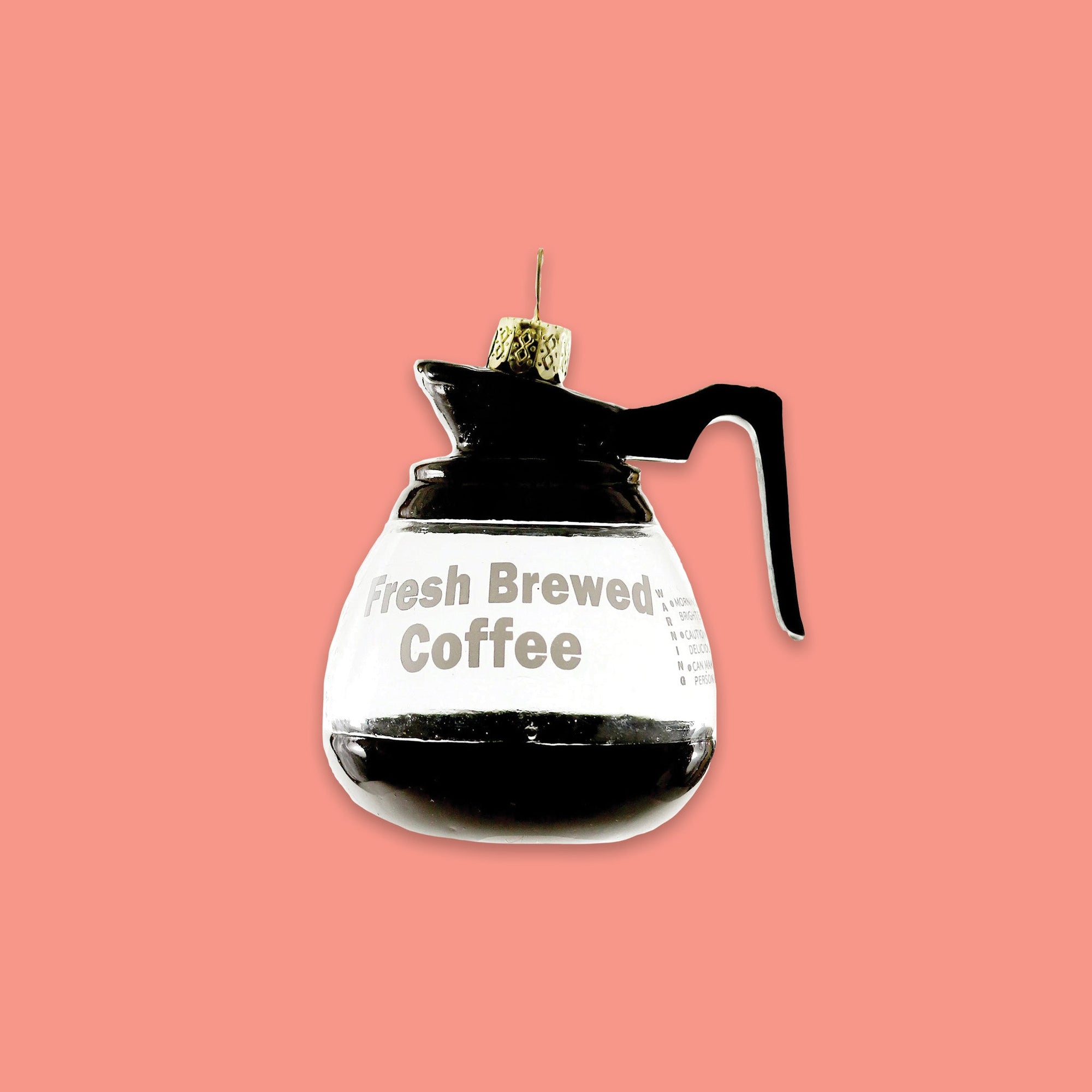On a coral pink background sits an ornament. This pot of coffee glass ornament says "Fresh Brewed Coffee." It looks like a real pot of coffee and there is coffee inside.