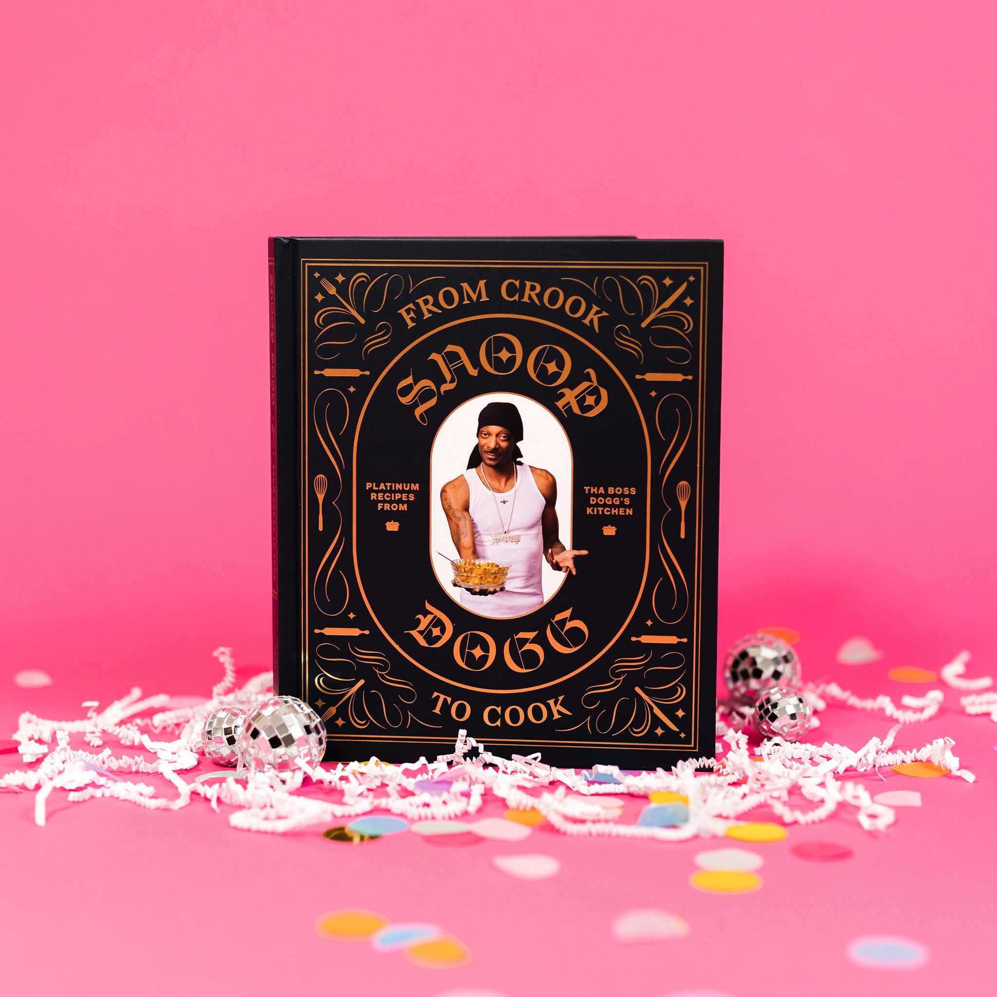 On a bubblegum pink background sits a book with white crinkle and big, colorful confetti scattered around. There are mini disco balls. This black cookbook has orange illustrations of kitchen utensils and a picture of Snoop Dogg holding a bowl of food. It says "FROM CROOK TO COOK SNOOP DOGG" and "PLATINUM RECIPES FROM THA BOSS DOGG'S KITCHEN."