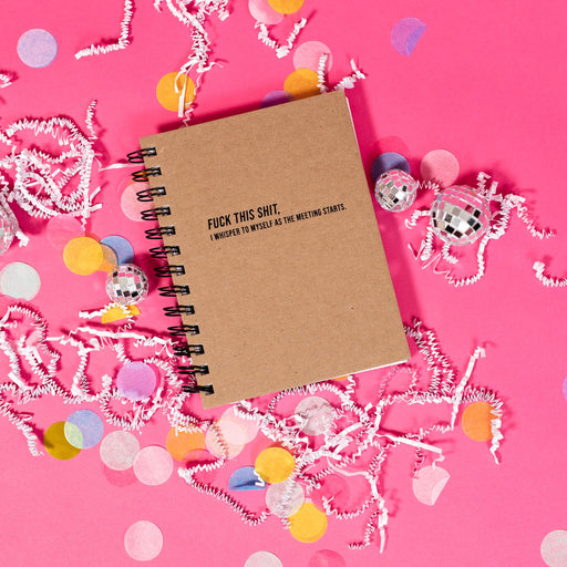 On a hot pink background sits a spiral notebook with white crinkle and big, colorful confetti scattered around. There are mini disco balls. This kraft notebook says "FUCK THIS SHIT, I WHISPER TO MYSELF AS THE MEETING STARTS." in black all caps font.