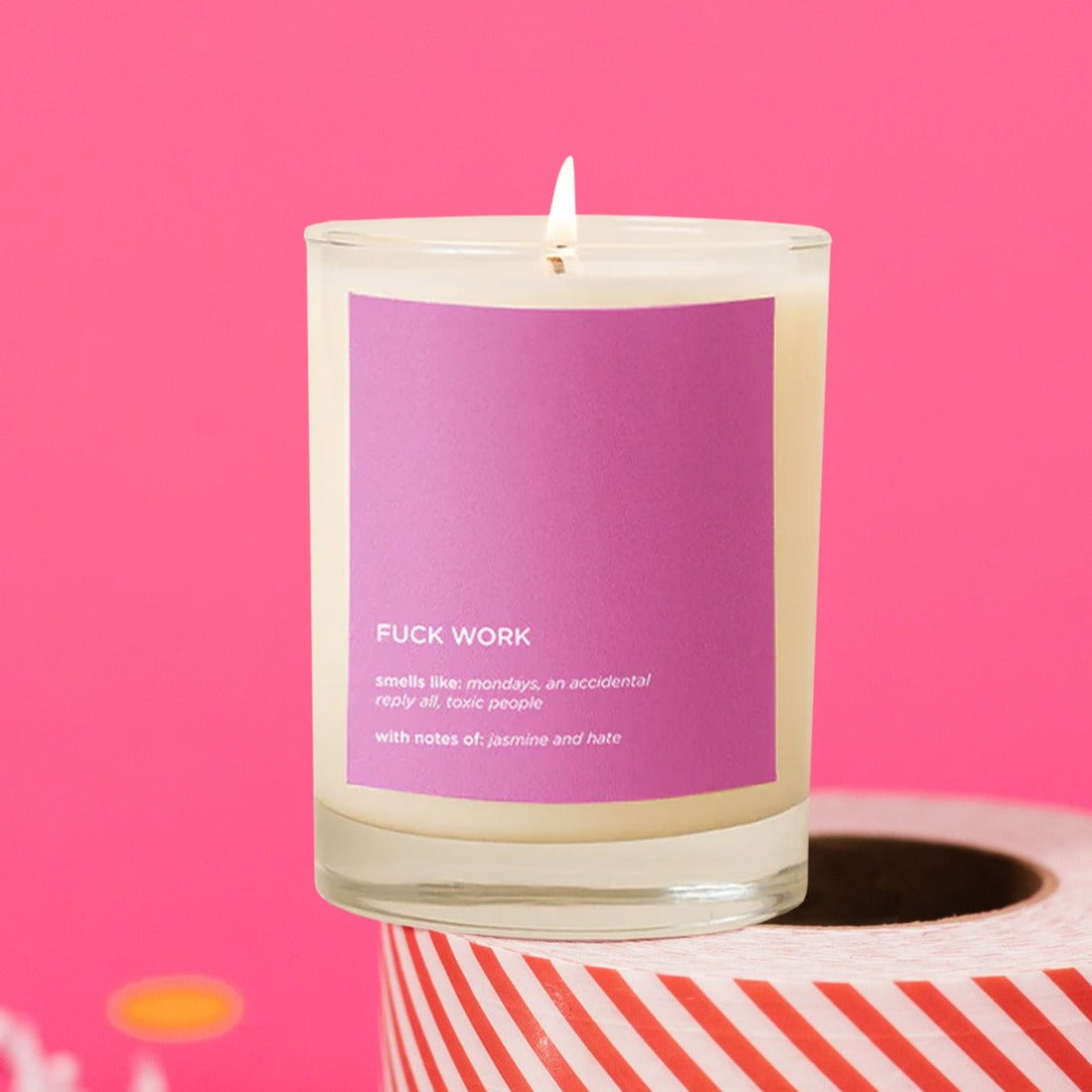 On a hot pink background sits a candle atop a roll of red and white striped packing tape. There are white crinkle and big, colorful confetti scattered around. The picture is a close-up of a glass candle that is lit and has a hot pink label on the front. It says "FUCK WORK," "smells like: mondays, an accidental reply all, toxic people," "with notes of: jasmine and hate." It is in a white, block font."