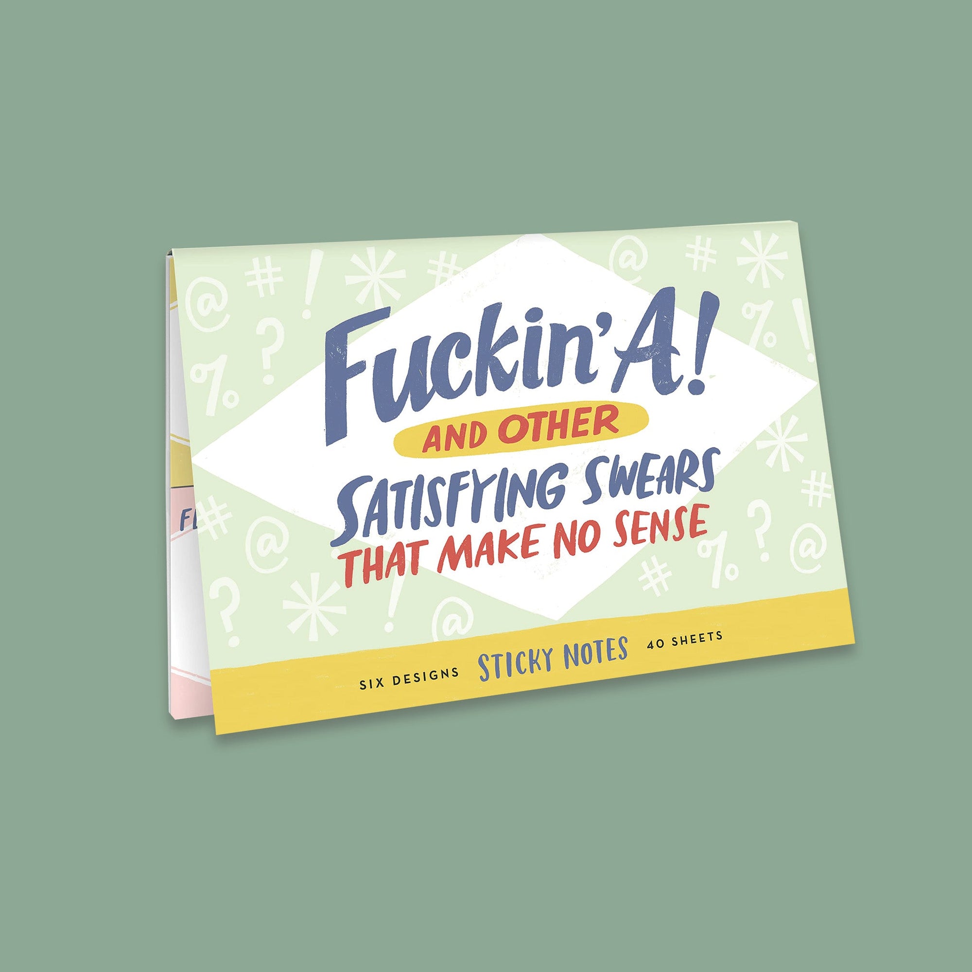 On a moss green background sits a packet. This is a close-up of a light green packet of sticky notes that has white illustrations of typing symbols. It says "Fuckin' A! AND OTHER SATISFYING SWEARS THAT MAKE NO SENSE" in handwritten lettering. The colors of the lettering are navy, light yellow, and white. On the bottom it says "SIX DESIGNS STICKY NOTES 40 SHEETS" in navy, handwritten lettering.