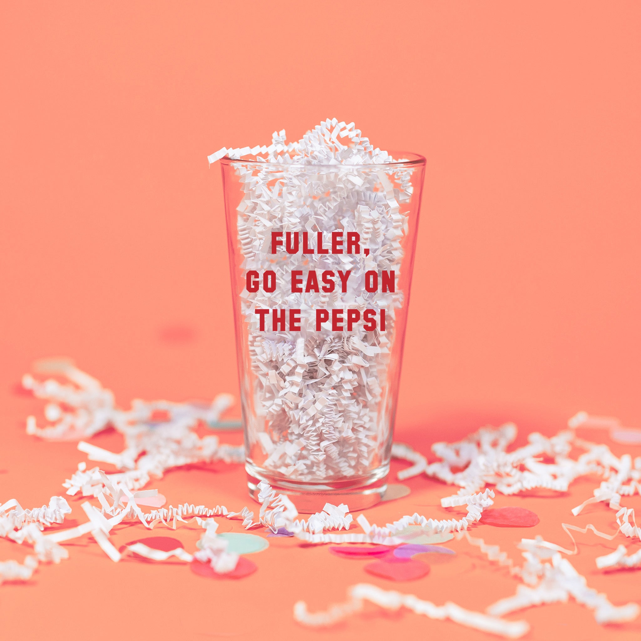 On a pink coral background sits a pint glass with white crinkle and big, colorful confetti scattered around. This Home Alone Inspired pint glass has red collegiate lettering and it says "FULLER, GO EASY ON THE PEPSI." There is white crinkle inside the glass.