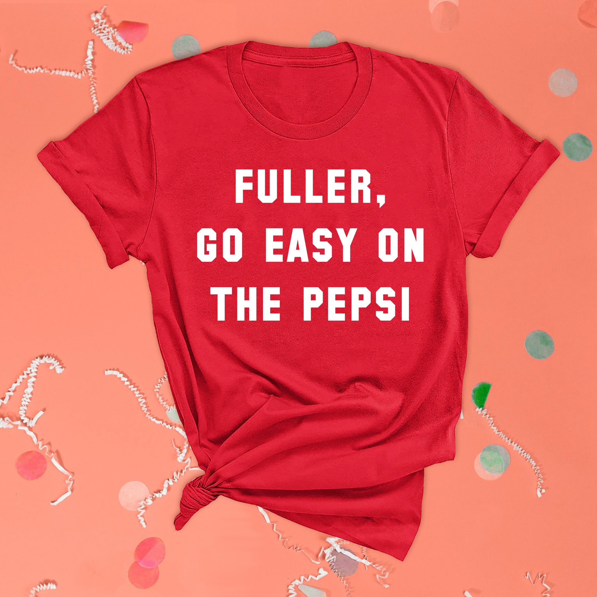 On a pink coral background sits the front of a t-shirt with white crinkle and big, colorful confetti scattered around. This Home Alone Inspired crewneck RED t-shirt has white collegiate lettering and it says "FULLER, GO EASY ON THE PEPSI."