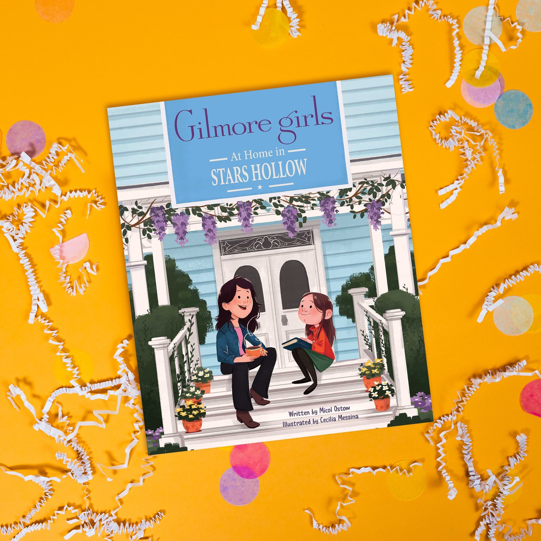 On a sunny mustard background sits a book with white crinkle and big, colorful confetti scattered around. This colorful Gilmore Girls inspired book is called "Gilmore girls At Home in STARS HOLLOW." It has an illustration of Lorelai and Rory sitting on the front steps of a house. Lorelai has a cup of coffee in her hand and Rory has a book on her lap. She is looking up at her mom.