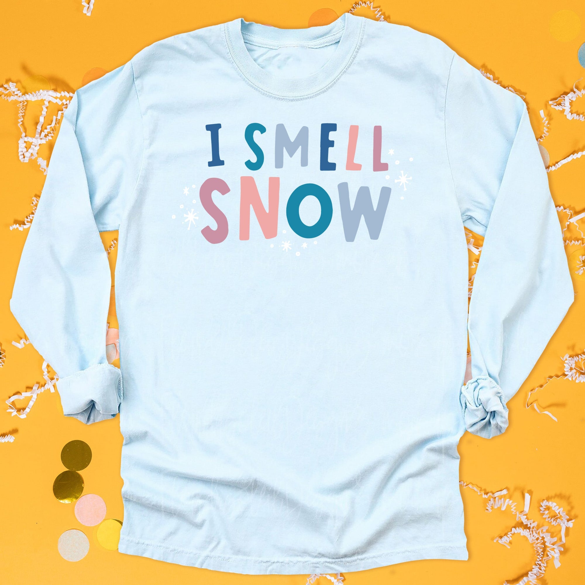 On a sunny mustard background sits a long sleeved tshirt with white crinkle and big, colorful confetti scattered around. This Gilmore Girls inspired tee is light blue with colorful lettering and snow scattered around them. It says "I SMELL SNOW" in all caps, handwritten block font. The colors are navy, teal, grey, coral, and rose.