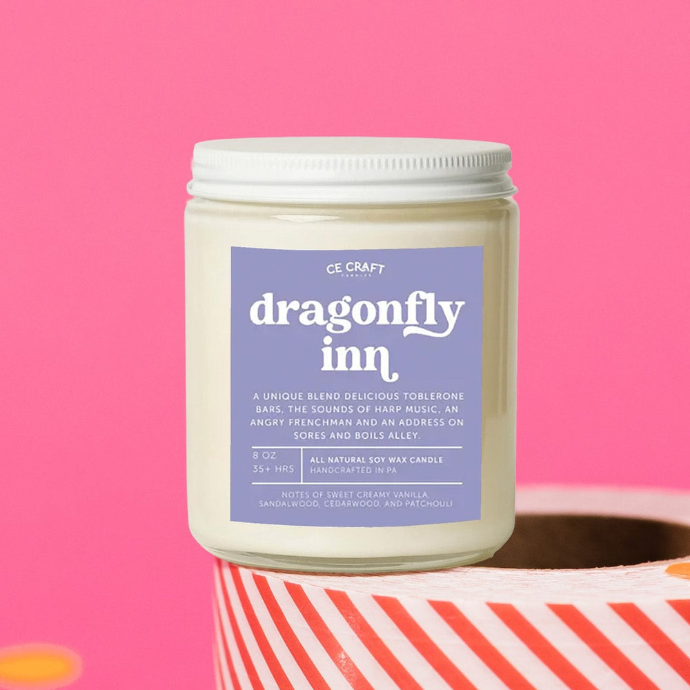 On a hot pink background sits a candle on a red and white packing tape with colorful confetti and white crinkle scattered around. It is has a lavender label with white text. This Dragonfly Inn Candle has notes of sweet creamy vanilla, sandalwood, and patchouli & is the perfect scent for your die hard Gilmore Girls Fan! This standard sized 8 oz jar has a burn time of approximately 35+ hours, perfect for bathrooms, dressers, countertops, and nightstands.