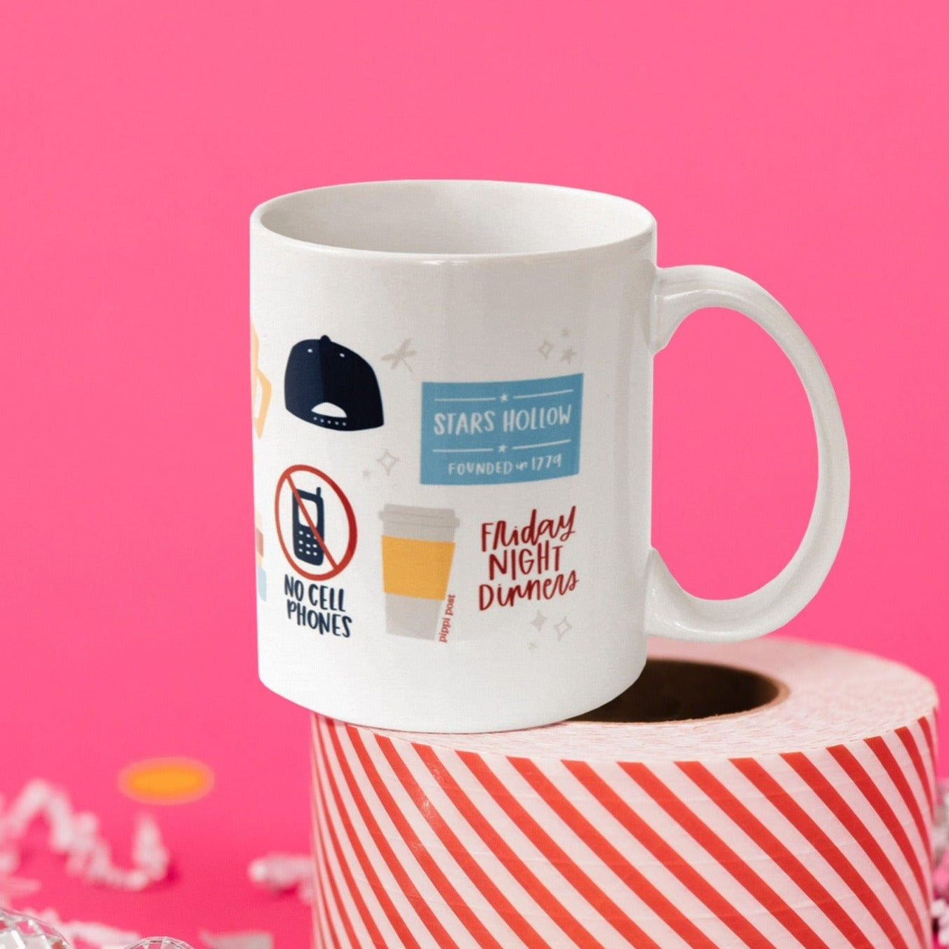 On a hot pink background sits a mug atop a red and white striped roll of packing tape with white crinkle and big, colorful confetti scattered around. There are mini disco balls. This Gilmore Girls inspired white mug has colorful illustrations from the show. It says "STARS HOLLOW FOUNDED in 1774." There is a navy backwards cap, a to-go coffee cup, a cell phone that has red circle and slash on top that says "NO CELL PHONES" and it also says "Friday NIGHT Dinners."