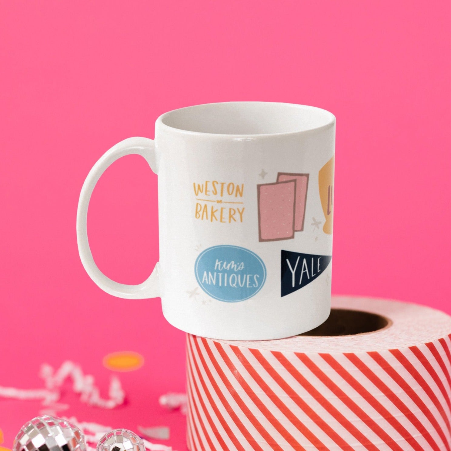 On a hot pink background sits a mug atop a red and white striped roll of packing tape with white confetti and big, colorful confetti scattered. There are mini disco balls. This Gilmore Girls inspired white mug has colorful illustrations from the show. It says "WESTON BAKERY" and "Kim's ANTIQUES." There are two pink menus and a navy school banner that says "YALE."