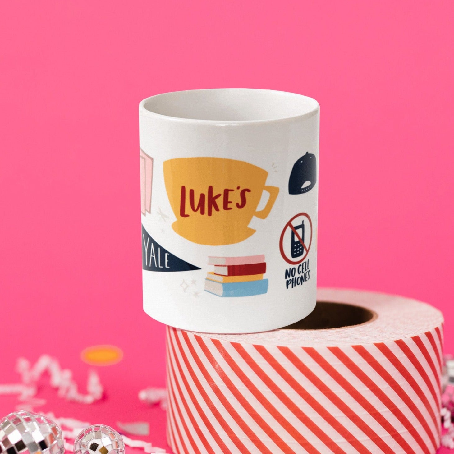 On a hot pink background sits a mug atop a red and white striped roll of packing tape with white crinkle and big, colorful confetti scattered around. There are mini disco balls. This Gilmore Girls inspired white mug has colorful illustrations from the show. It has a coffee mug that says "Luke's." There are books stacked, a cell phone with a red circle and slash on top that says "NO CELL PHONES." There is a navy school banner that says "YALE."