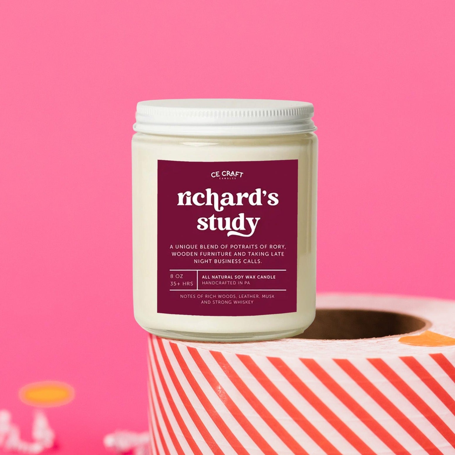 On a hot pink background sits a candle on a red and white packing tape with colorful confetti and white crinkle scattered around. It is has a maroon label with white text. This Richard's Study Candle has notes of rich woods, leather, musk & strong whiskey & is the perfect scent for your die hard Gilmore Girls Fan! Candle is 8 oz and burns 35 hours.