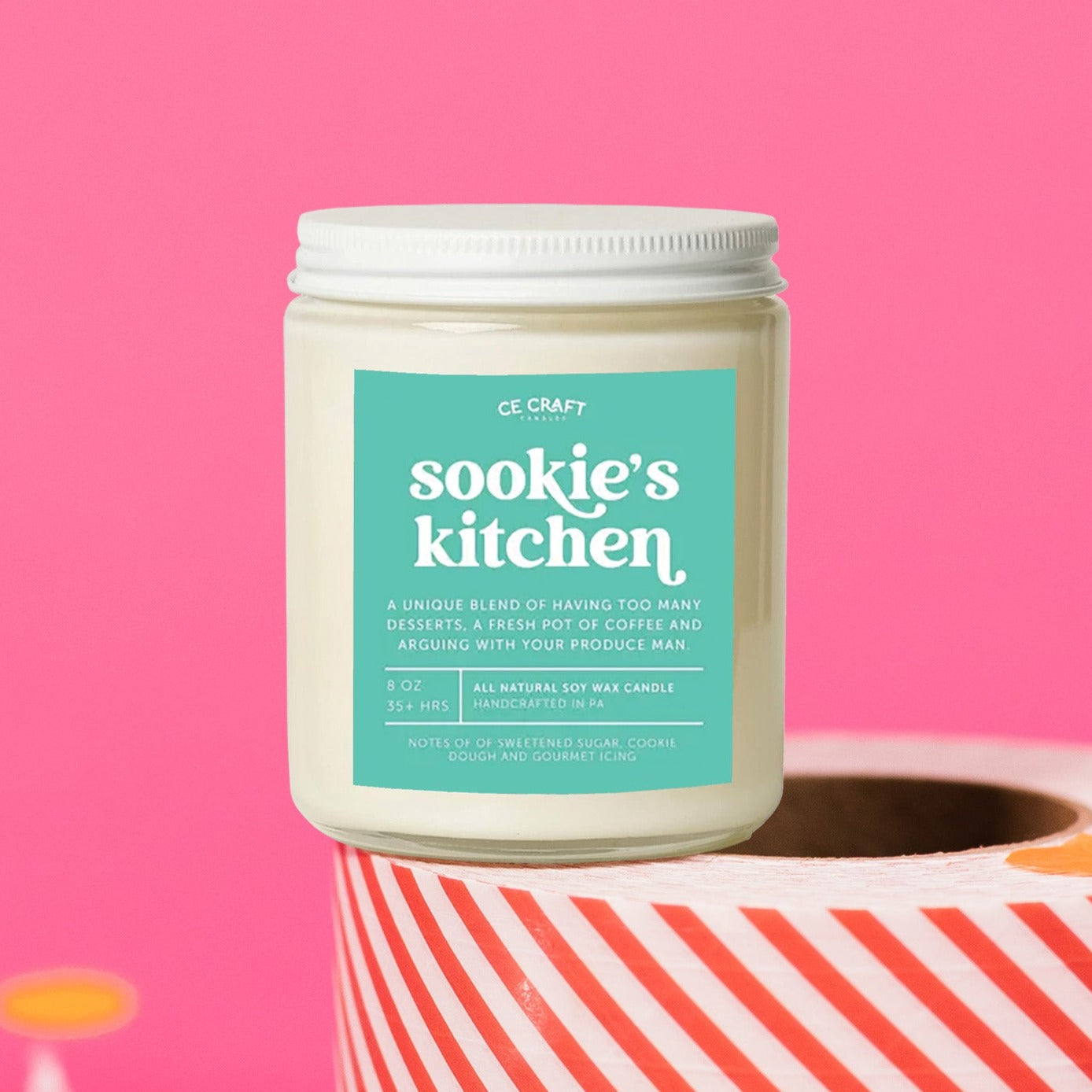 On a hot pink background sits a candle on a red and white packing tape with colorful confetti and white crinkle scattered around. It is has a mint green label with white text. This Sookie's Kitchen Candle has notes of sweetened sugar, cookie dough and gourmet icing and is the perfect scent for your die hard Gilmore Girls Fan! Candle is 8 oz and burns 35 hours.