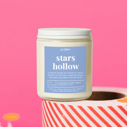 On a hot pink background sits a candle on a red and white packing tape with colorful confetti and white crinkle scattered around. It is has an orange label with white text. This Stars Hollow Fall Festival Candle has notes of creamy vanilla, fluffy marshmallows and pumpkin pie and is the perfect scent for your die hard Gilmore Girls Fan! Candle is 8 oz and burns 35 hours.