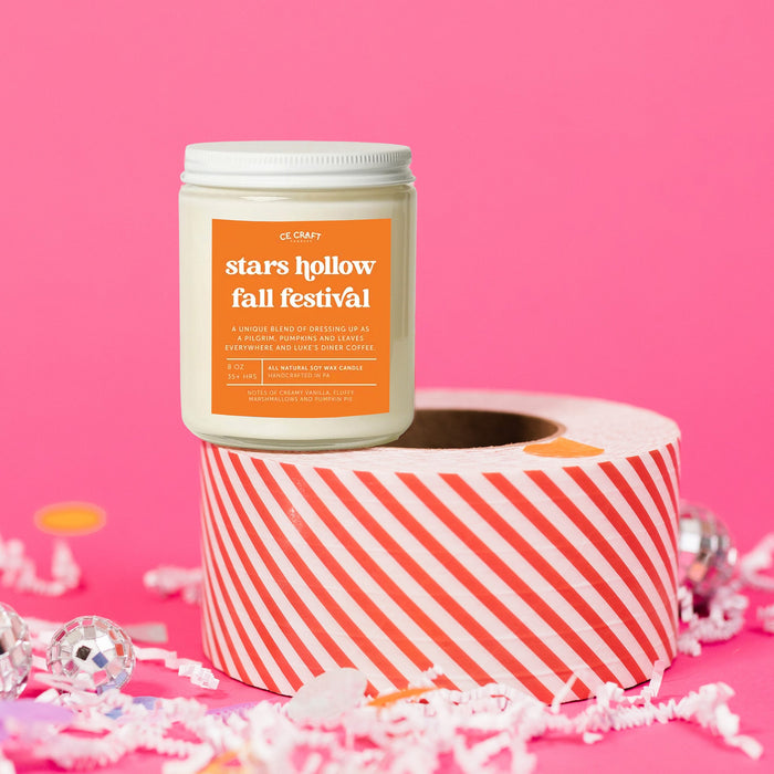 On a hot pink background sits a candle on a red and white packing tape with colorful confetti and white crinkle scattered around. It is has an orange label with white text. This Stars Hollow Fall Festival Candle has notes of creamy vanilla, fluffy marshmallows and pumpkin pie and is the perfect scent for your die hard Gilmore Girls Fan! Candle is 8 oz and burns 35 hours.