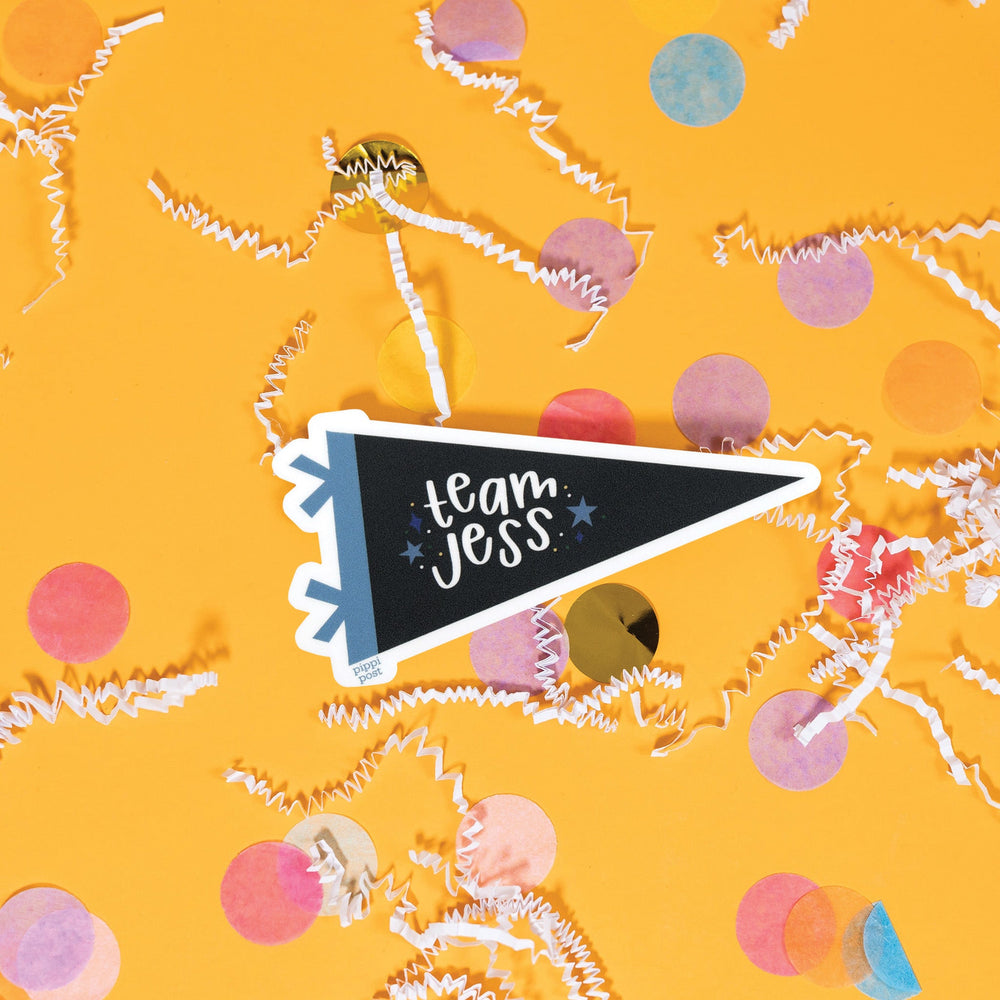 On a sunny mustard background is a sticker with white crinkle and big, colorful confetti scattered around. This Gilmore girls inspired vintage banner shaped sticker is new blue and khaki and has navy and orange stars. It says "team logan" in white, lower case handwritten lettering. 3"x1.6"
