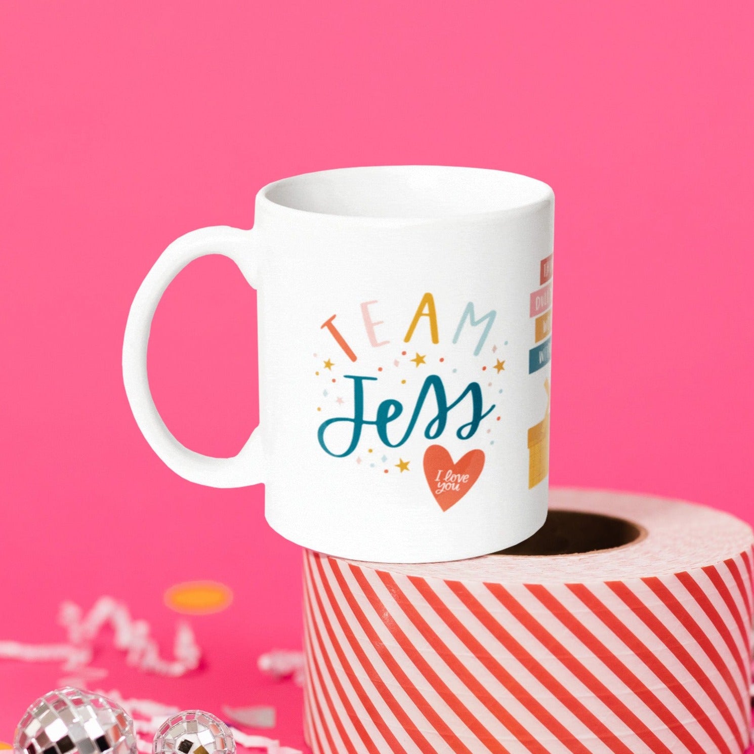 On a hot pink background sits a mug atop a red and white striped roll of packing tape with white crinkle and big, colorful confetti scattered around. There are mini disco balls. This Gilmore Girls inspired white mug says "TEAM Jess” and there are stars and colorful dots with a red heart at the bottom that says "I love you.”