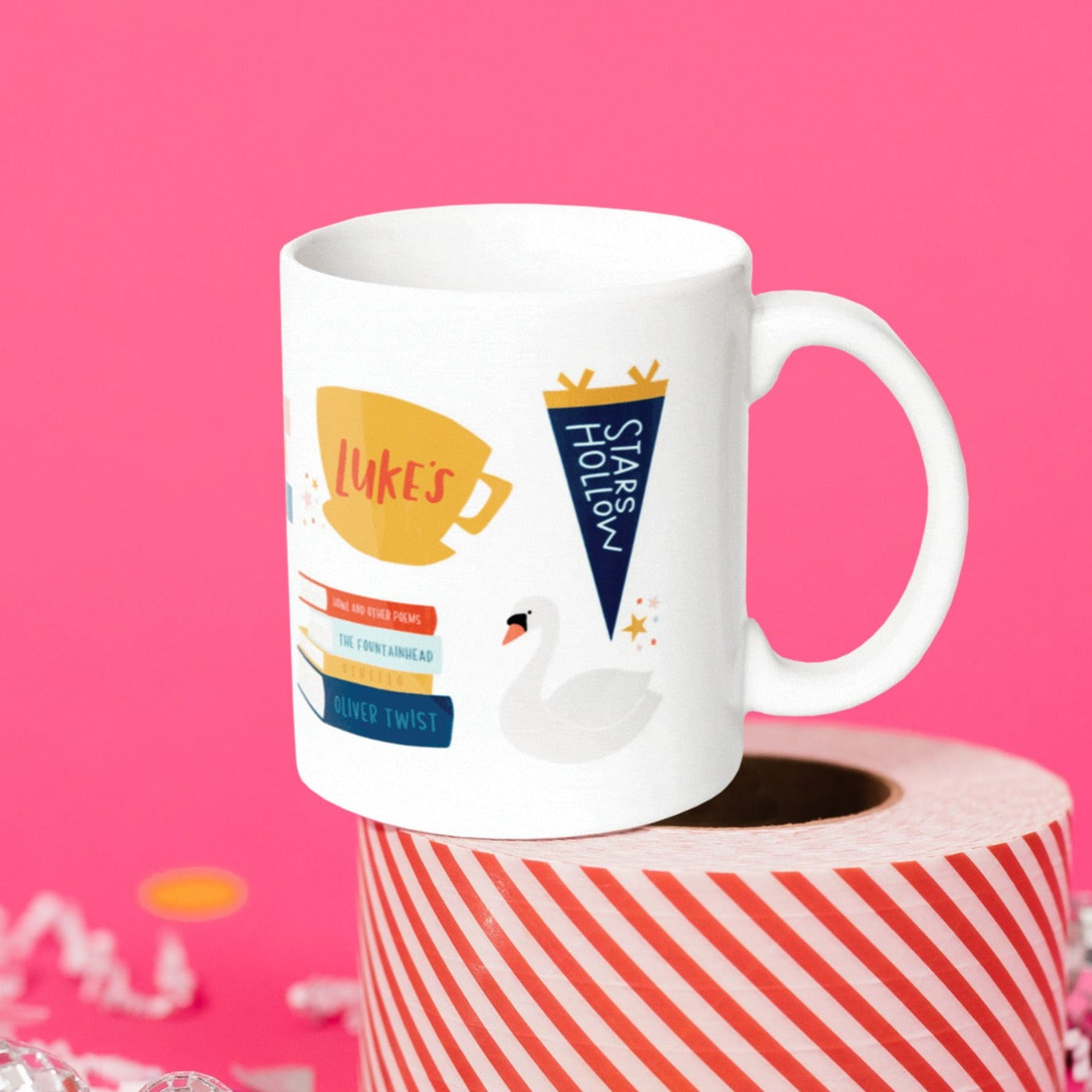 On a hot pink background sits a mug atop a red and white striped roll of packing tape with white crinkle and big, colorful confetti scattered around. There are mini disco balls. This Gilmore Girls inspired white mug has colorful illustrations from the show. It has a coffee mug that says “Luke's"and there are a stack of books, a swan, and a navy school banner that says "STARS HOLLOW.”