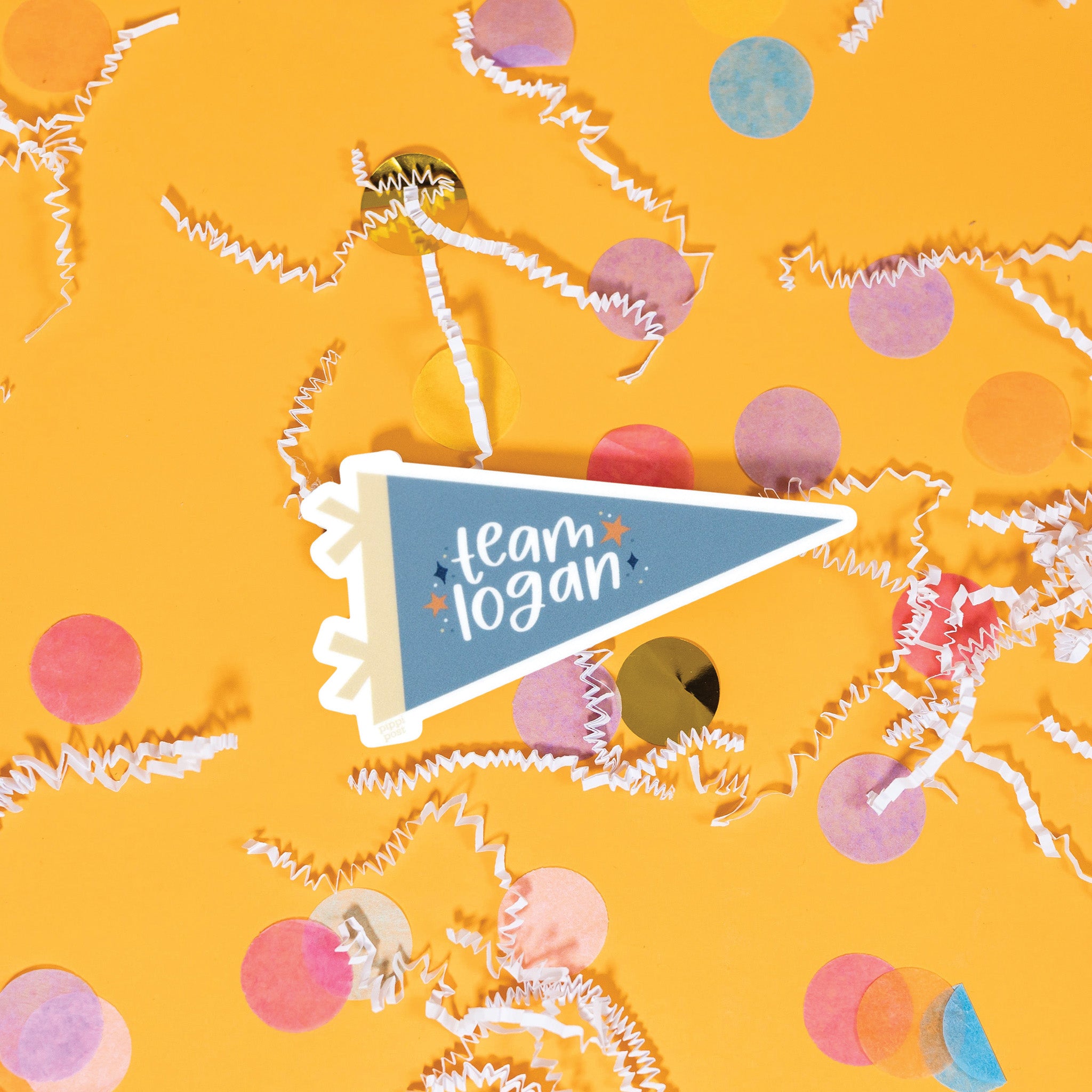 On a sunny mustard background is a sticker with white crinkle and big, colorful confetti scattered around. This Gilmore girls inspired vintage banner shaped sticker is new blue and khaki and has navy and orange stars. It says "team logan" in white, lower case handwritten lettering. 3"x1.6"