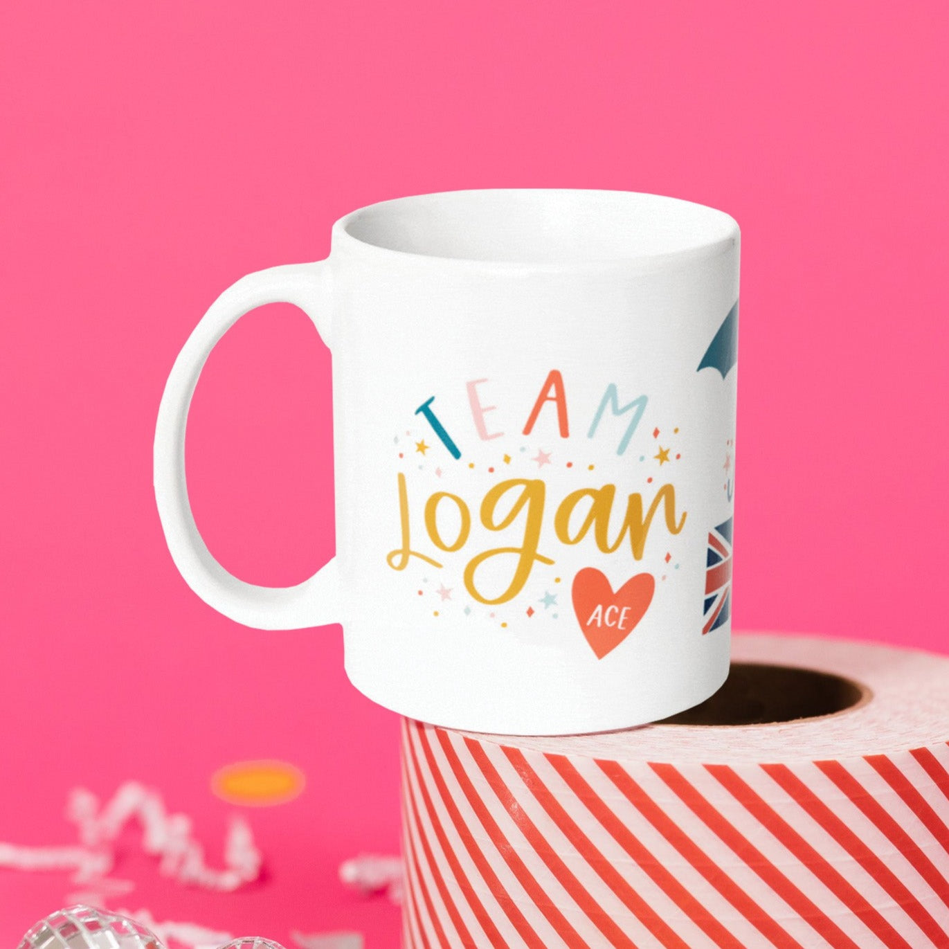 On a hot pink background sits a mug atop a red and white striped roll of packing tape with white crinkle and big, colorful confetti scattered around. There are mini disco balls. This Gilmore Girls inspired white mug says "TEAM Logan" in a colorful, handwritten lettering. There are stars and colorful dots and a red heart at the bottom that says "ACE" in white handwritten lettering.